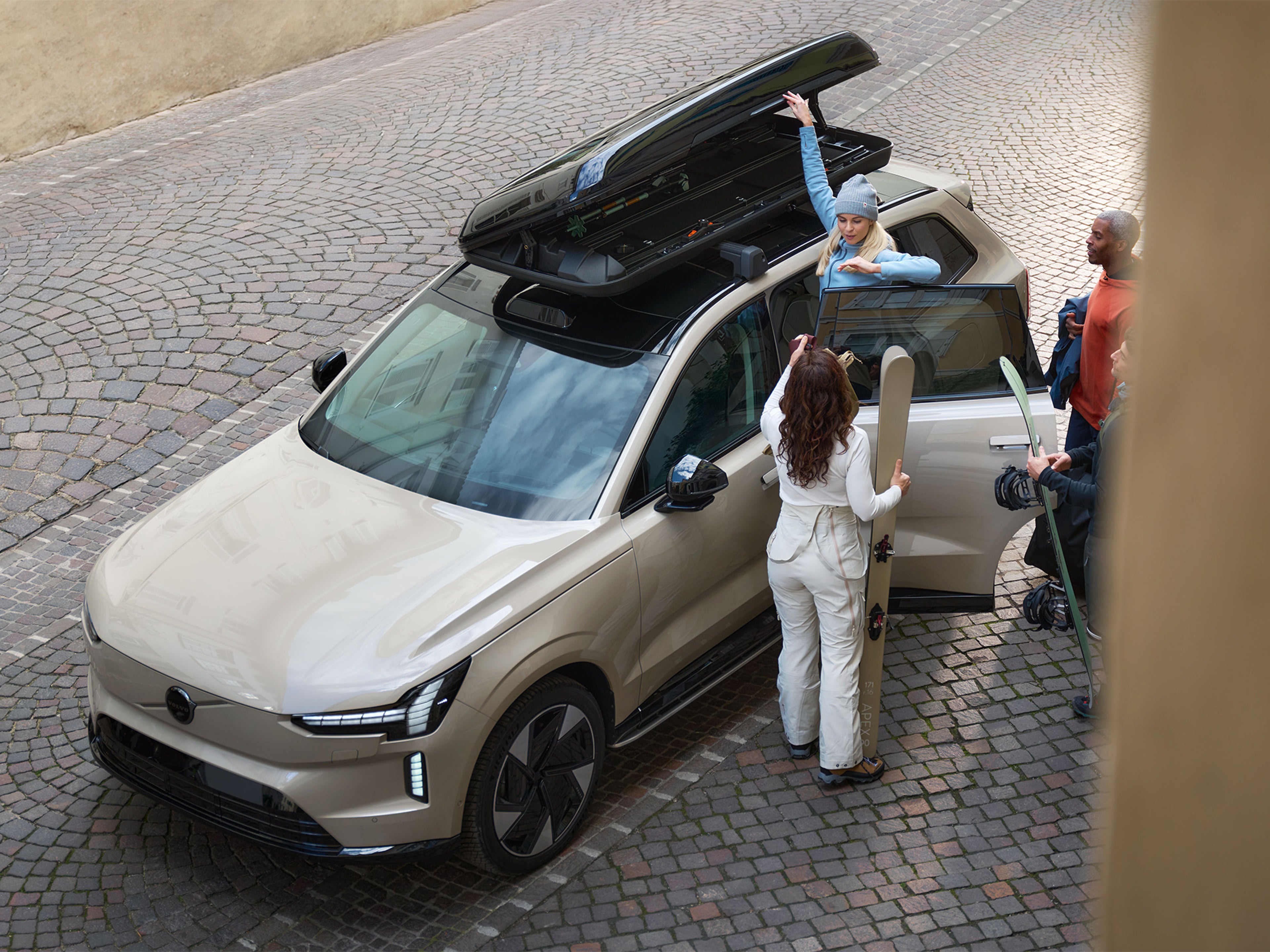 Dve ženski in dva moška stojijo ob Volvu EX90 in v strešni kovček zlagajo smučarsko opremo.