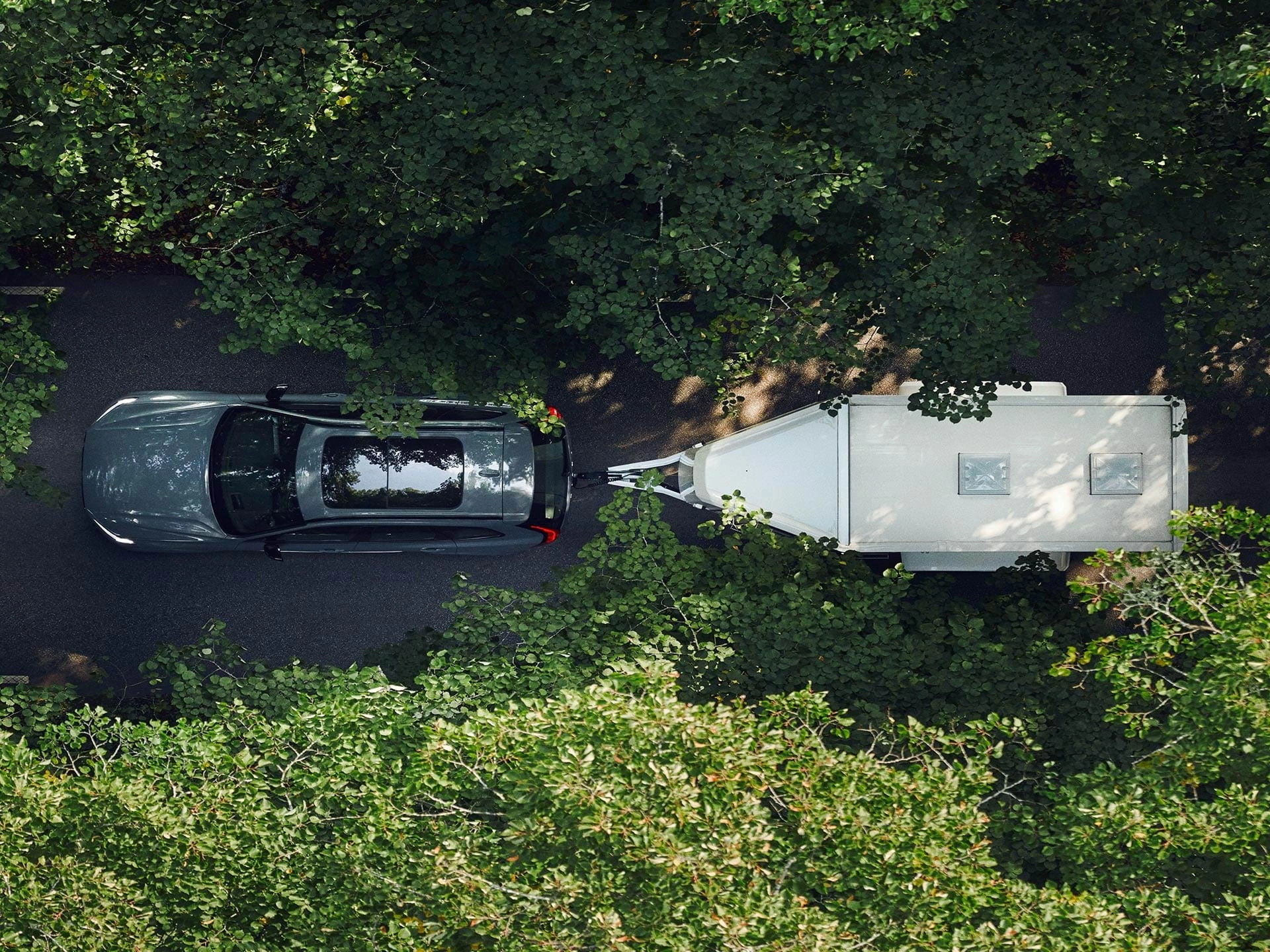 Perspectiva superior de um SUV Volvo a rebocar um atrelado branco numa estrada com folhas num dia de sol.