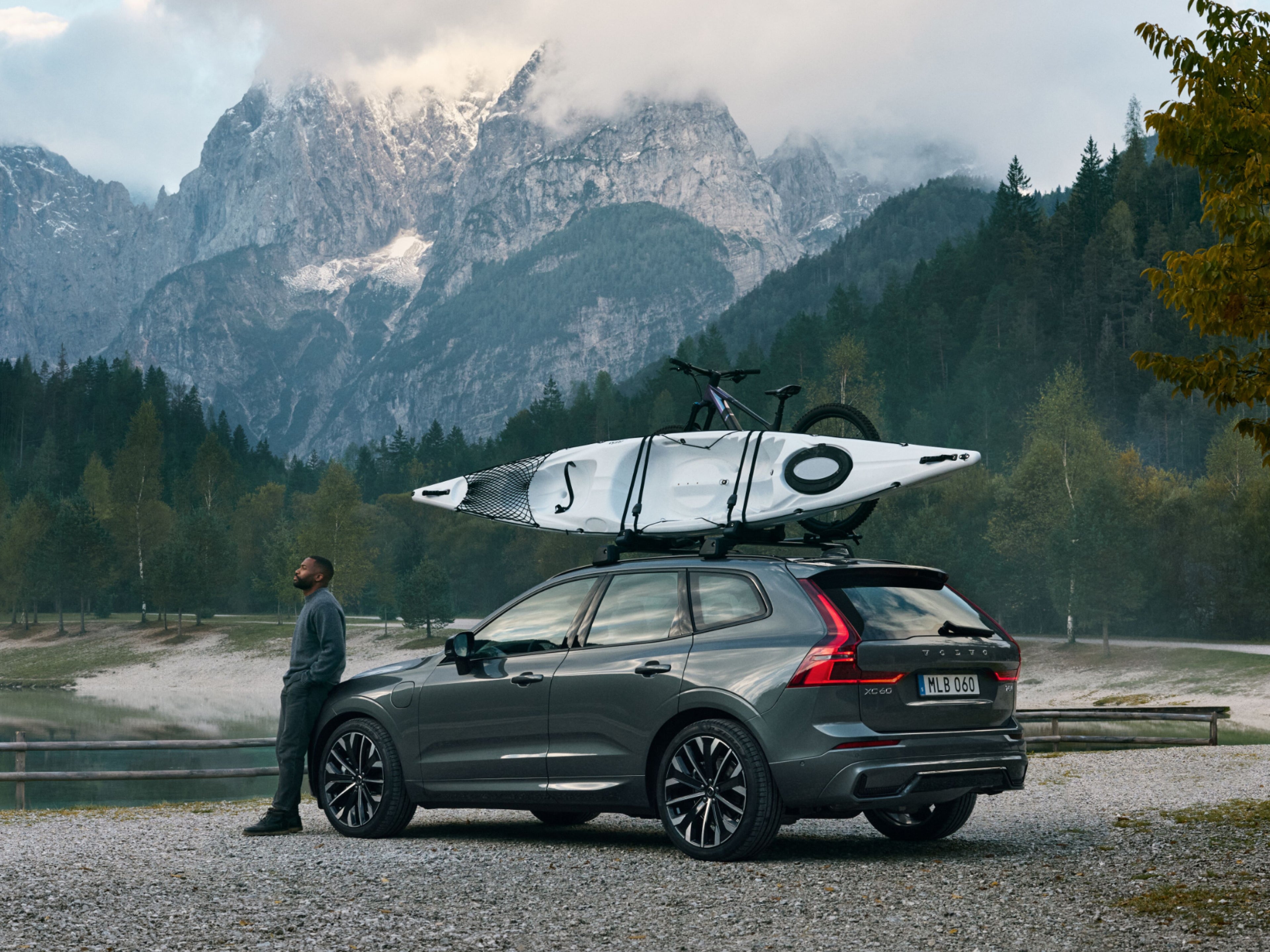 Widok z boku ciemnoszarego SUV-a EX60 zaparkowanego nad spokojnym jeziorem z górami w tle Na dachu zamontowany jest kajak i rower, a mężczyzna od niechcenia opiera się o przód samochodu.