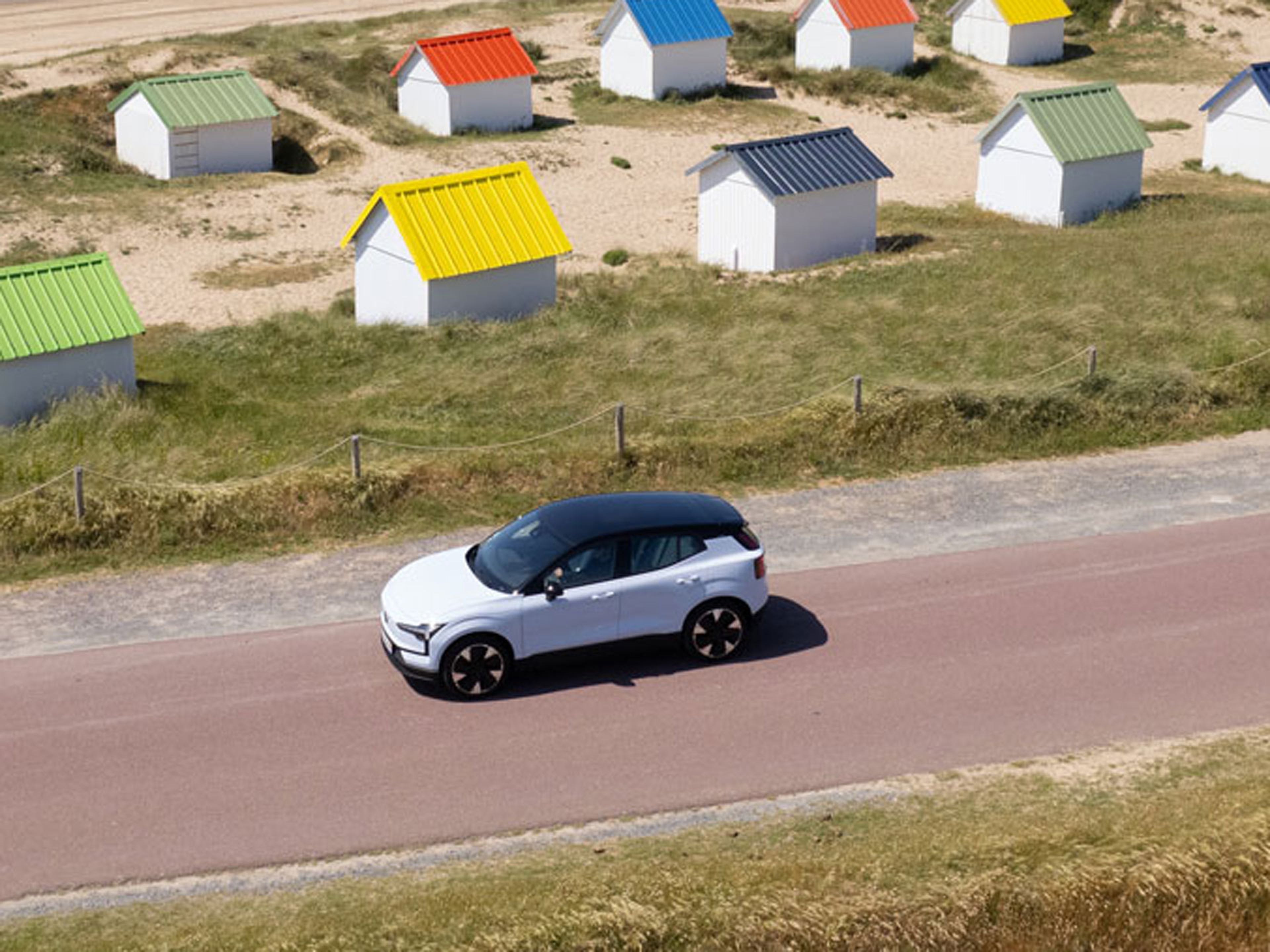 Volvo blanc sur route côtière, cabanes de plage colorées en arrière-plan près de la mer.