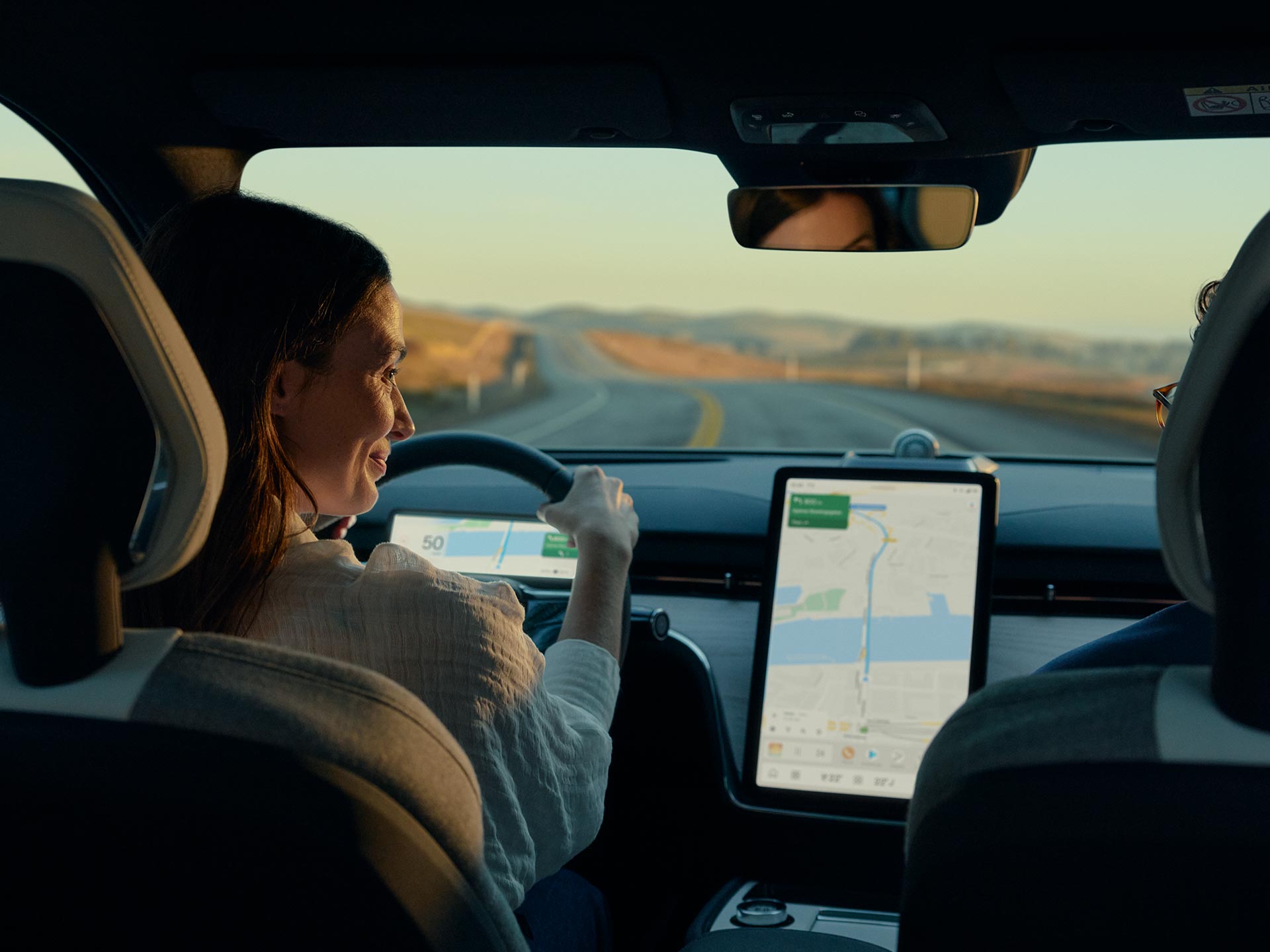 Жінка посміхається, дивлячись на великий яскравий центральний дисплей у своєму автомобілі Volvo, вигляд із заднього пасажирського сидіння.