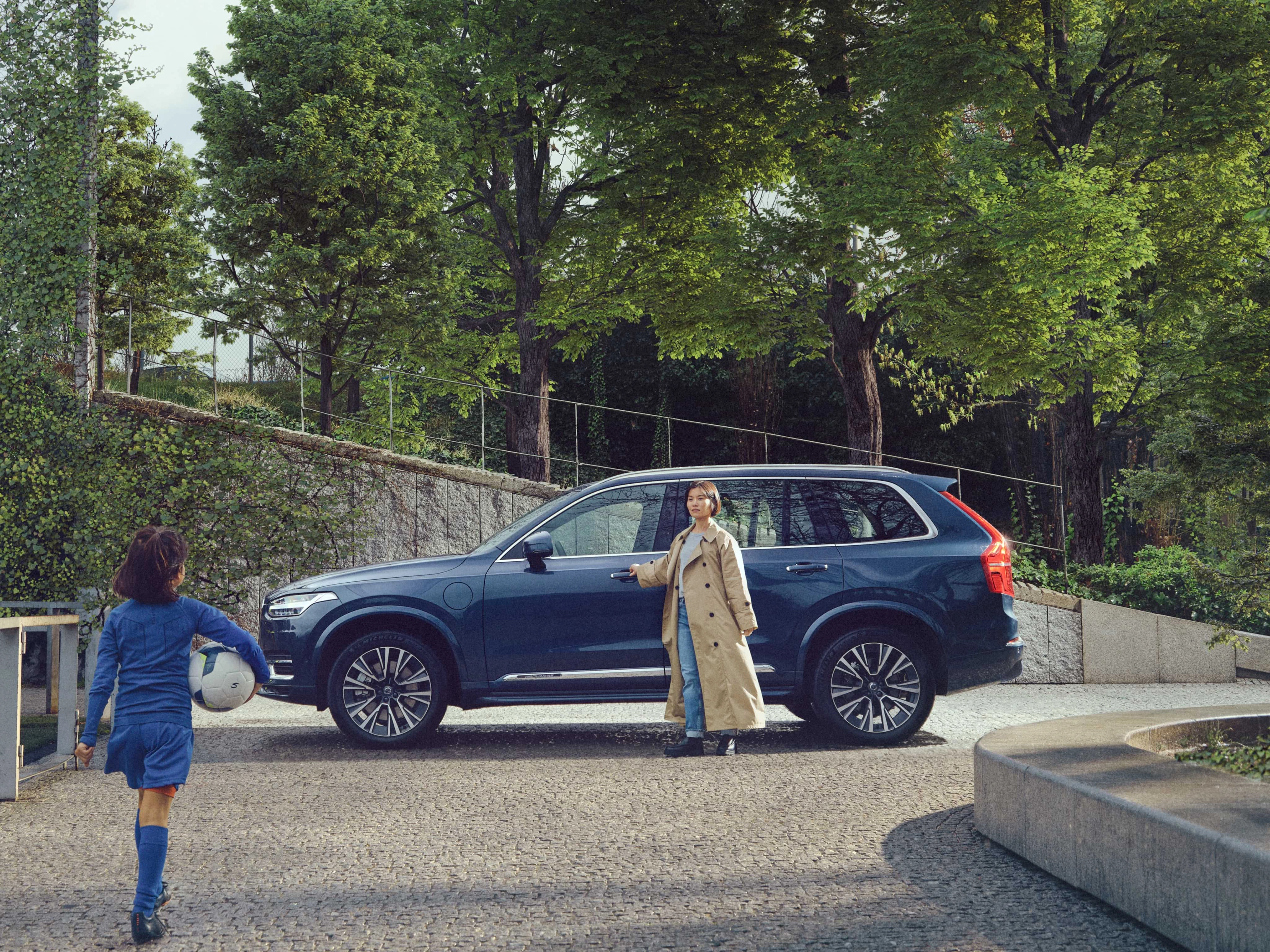Una madre permanece de pie junto a su Volvo XC90 Azul Denim mientras su hija se acerca, vestida para el entrenamiento de fútbol y con un balón en la mano.