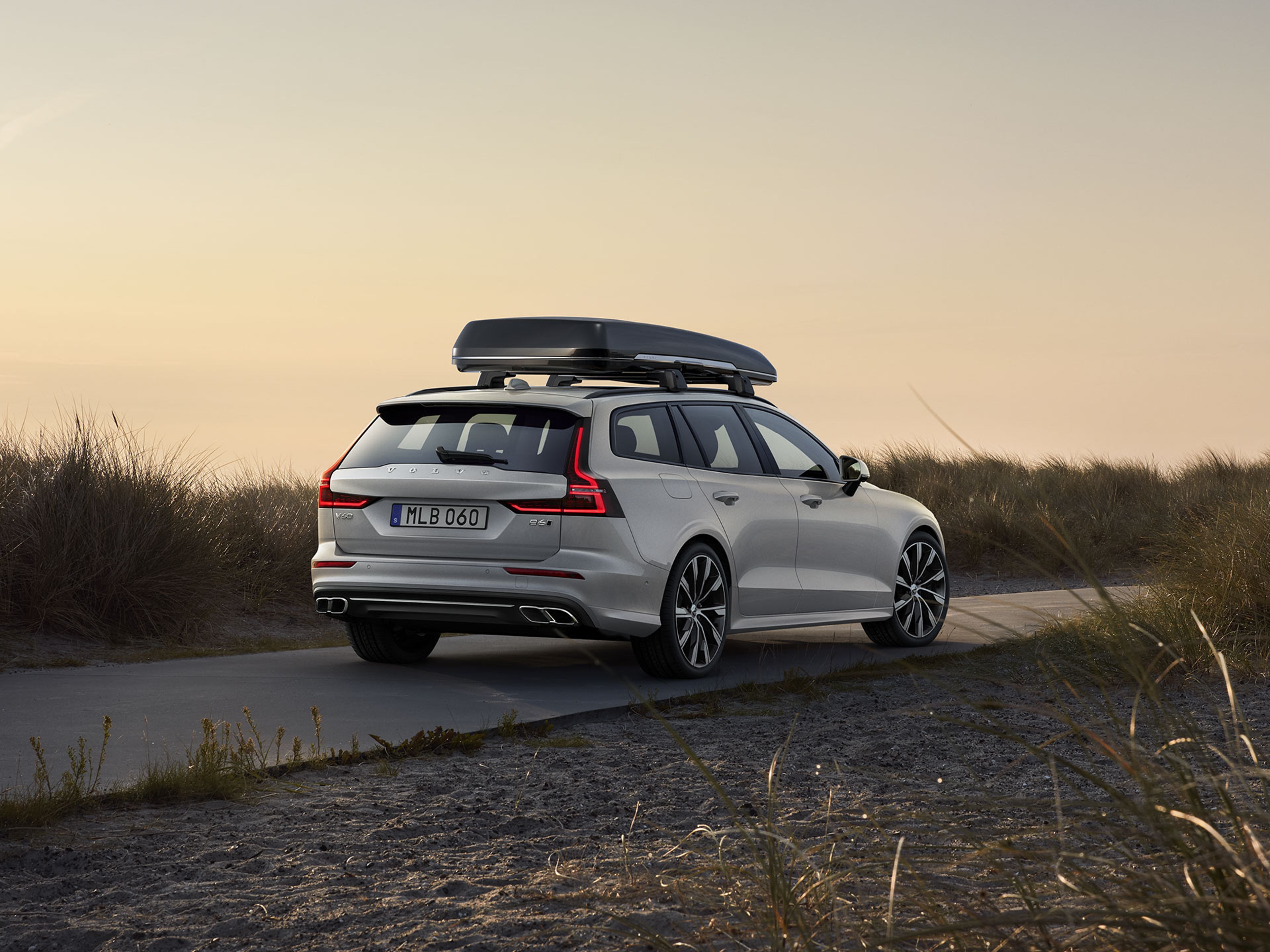 A white Volvo V60 wagon with a roof box accessory is parked on a coastal road at sunset, surrounded by sand dunes and tall grass under a clear sky.