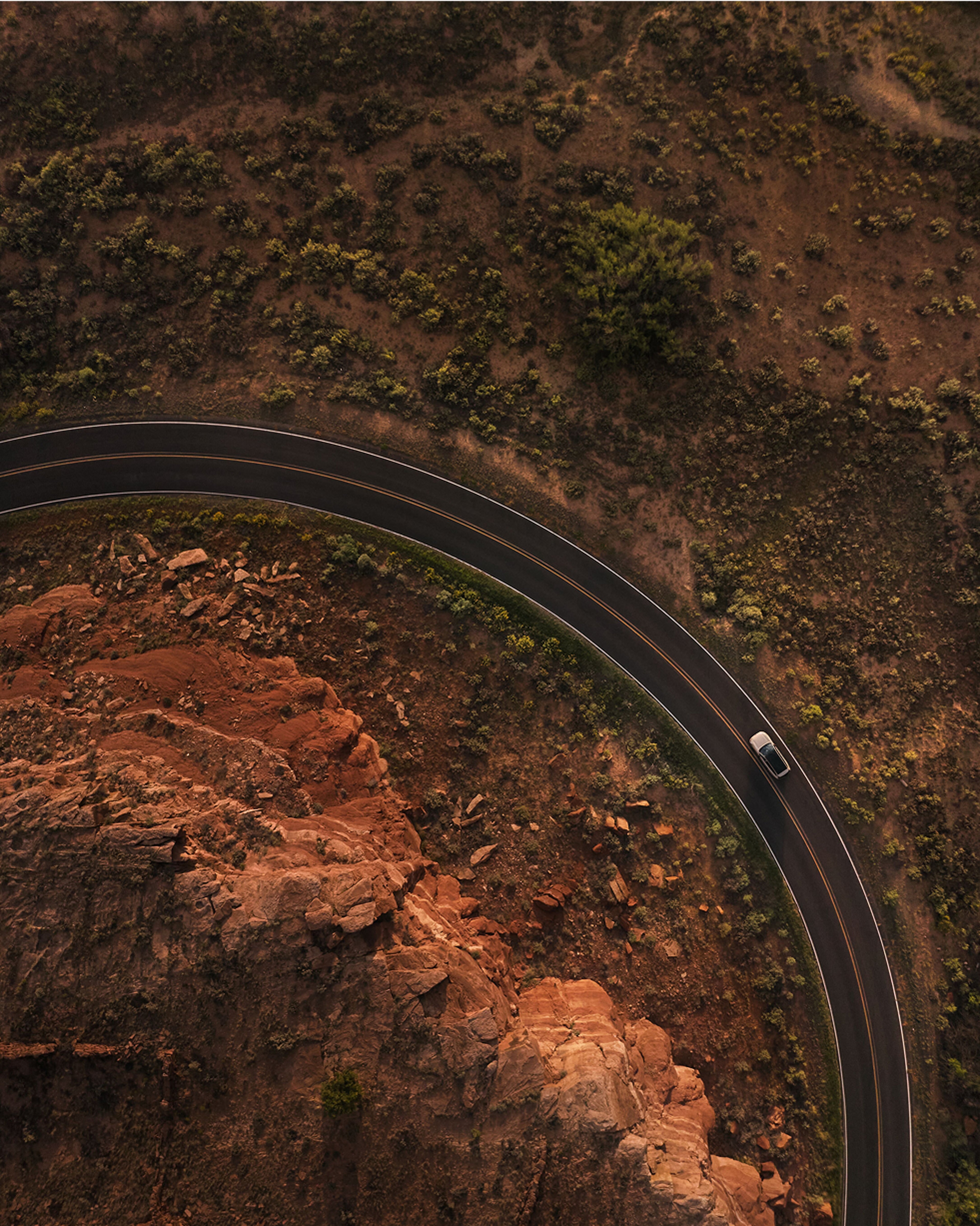 A Volvo EX90 drives along a winding road that curves sharply through a desert landscape, with red rocky cliffs on one side and sparse vegetation on the other, captured from above in warm, golden light. Component