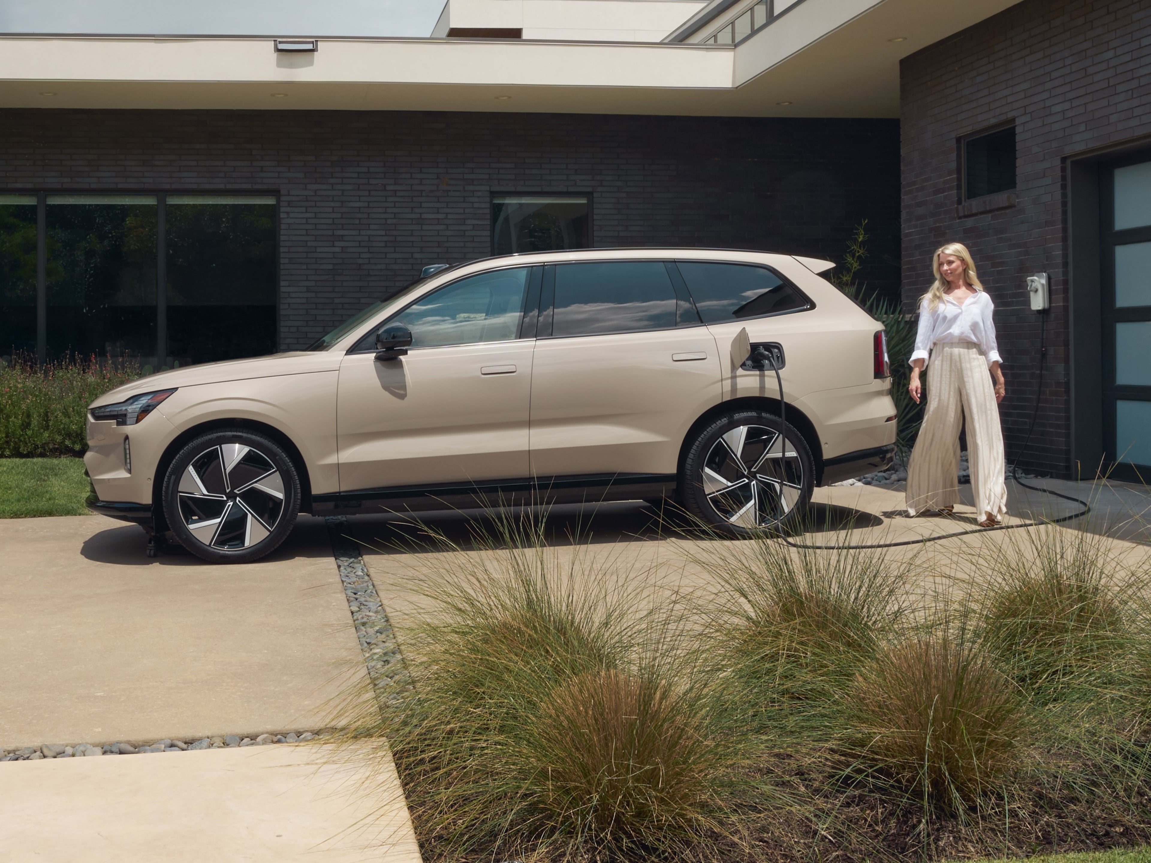 Side view of an electric Volvo car plugged into a home 
wallbox against a red home exterior.
