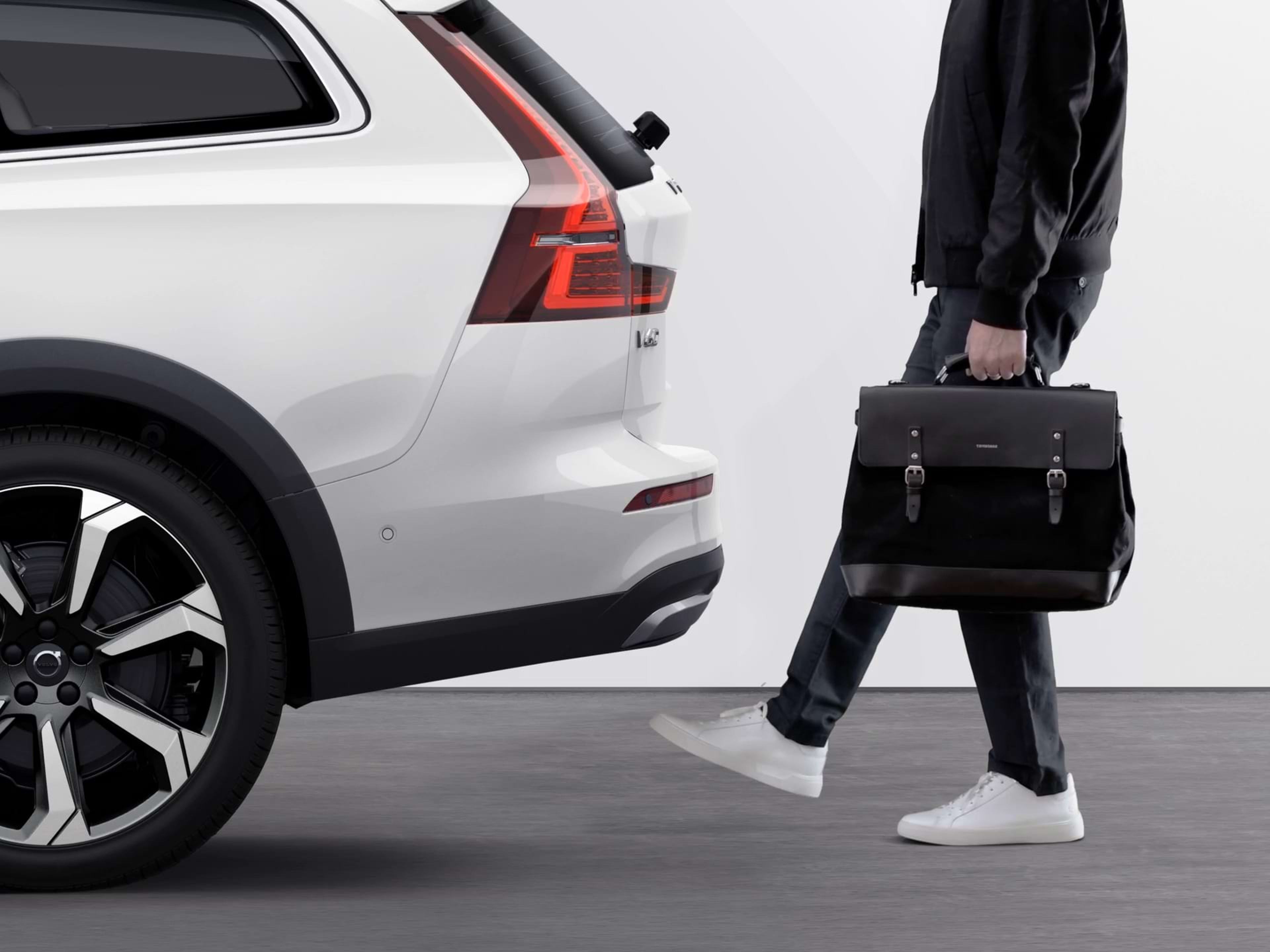 Un hombre que sujeta objetos con ambas manos pasa el pie por debajo del parachoques de un vehículo Volvo para abrir el portón trasero eléctrico.