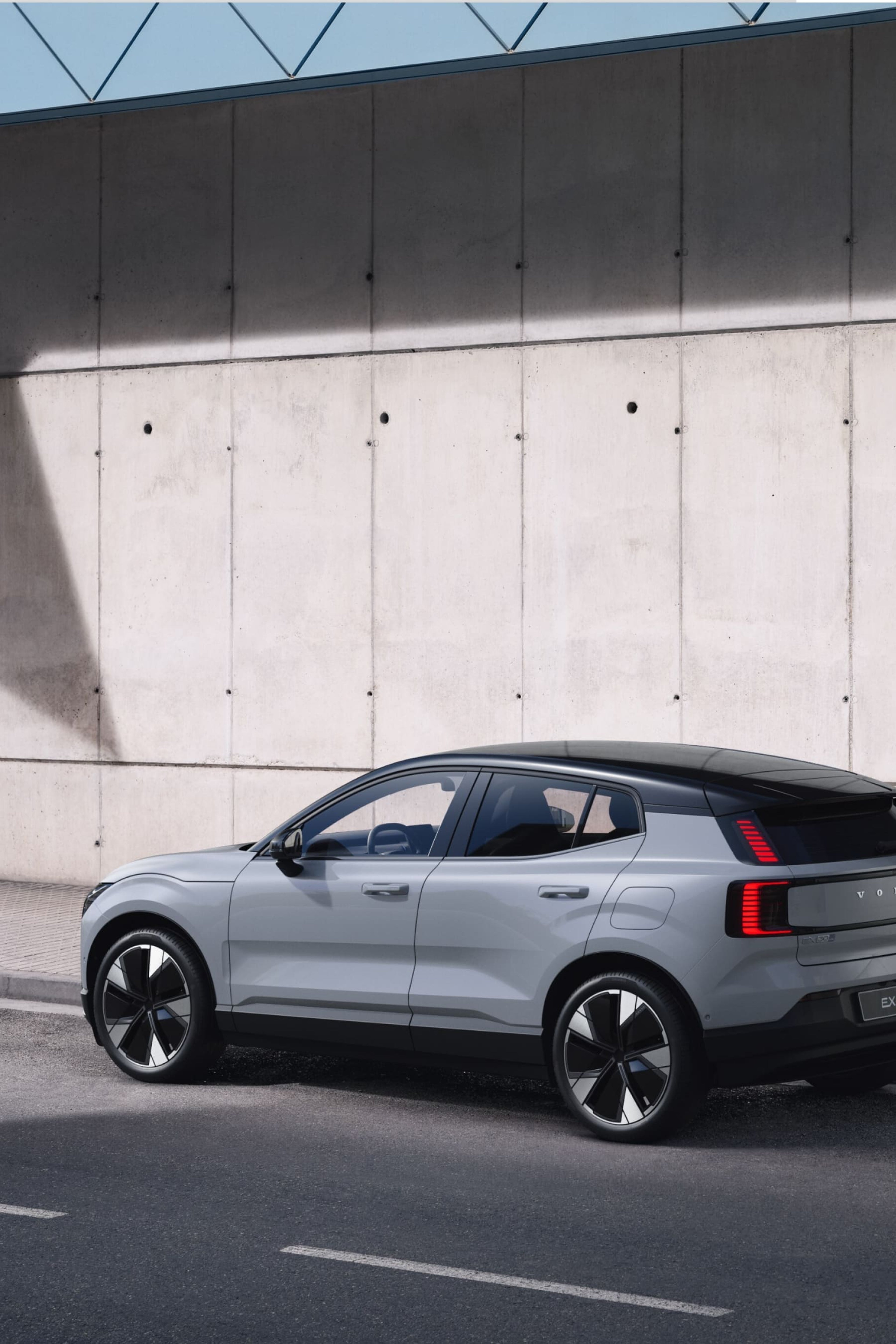 Une Volvo EX30 en Vapour Grey garée le long d'un mur en béton. La voiture a un toit noir et des feux arrière distinctifs.
