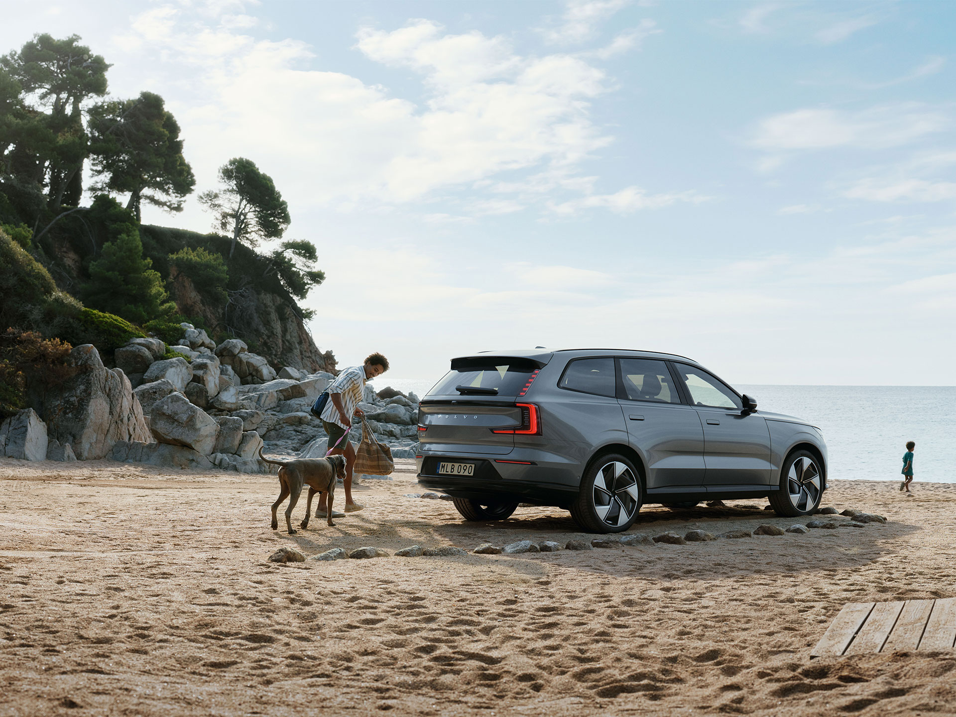 Achteraanzicht van een volledig elektrische Volvo EX90 op het strand met een man, een hond en twee kinderen die naar de zee rennen.