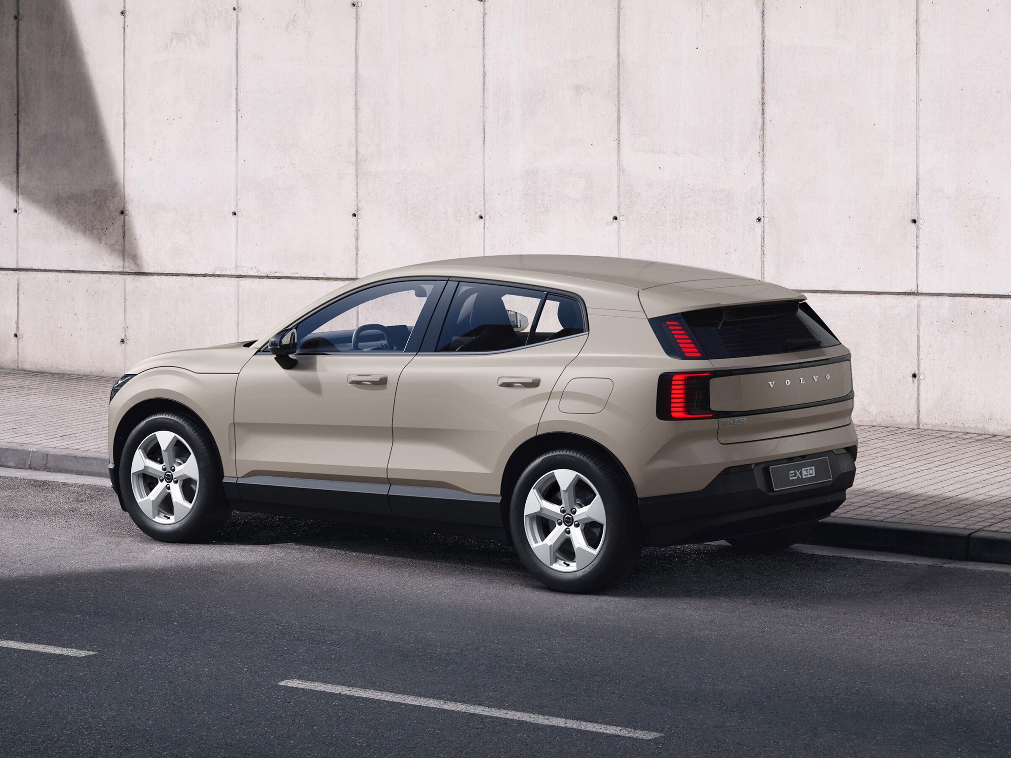 Sand dune -värinen Volvo EX30 Essential täyssähköinen katumaasturi pysäköitynä tien varteen. Taustalla jalkakäytävä ja betoniseinä.