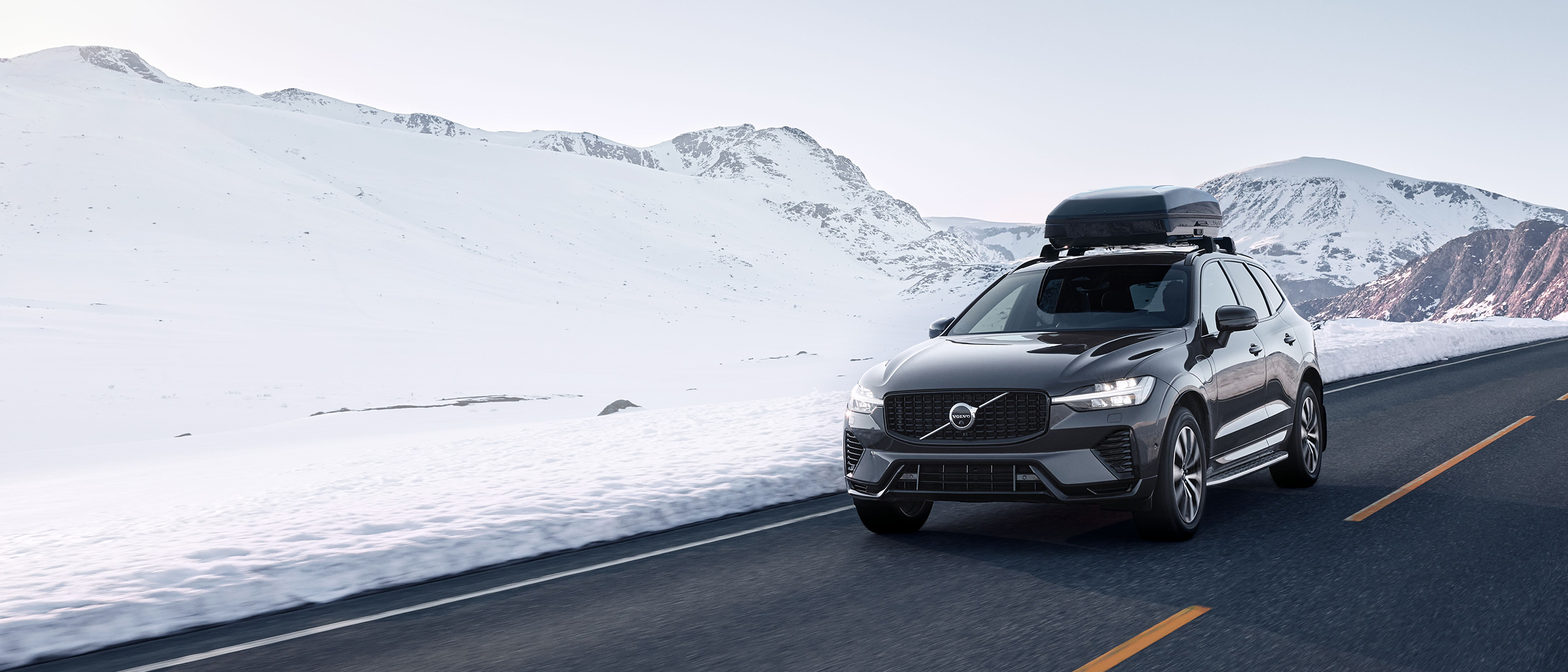 Platinum Grey -värinen Volvo XC60, jolla on kattolaatikko kinnitetty katolle, ajaa talvisessa vuoristomaisemassa.