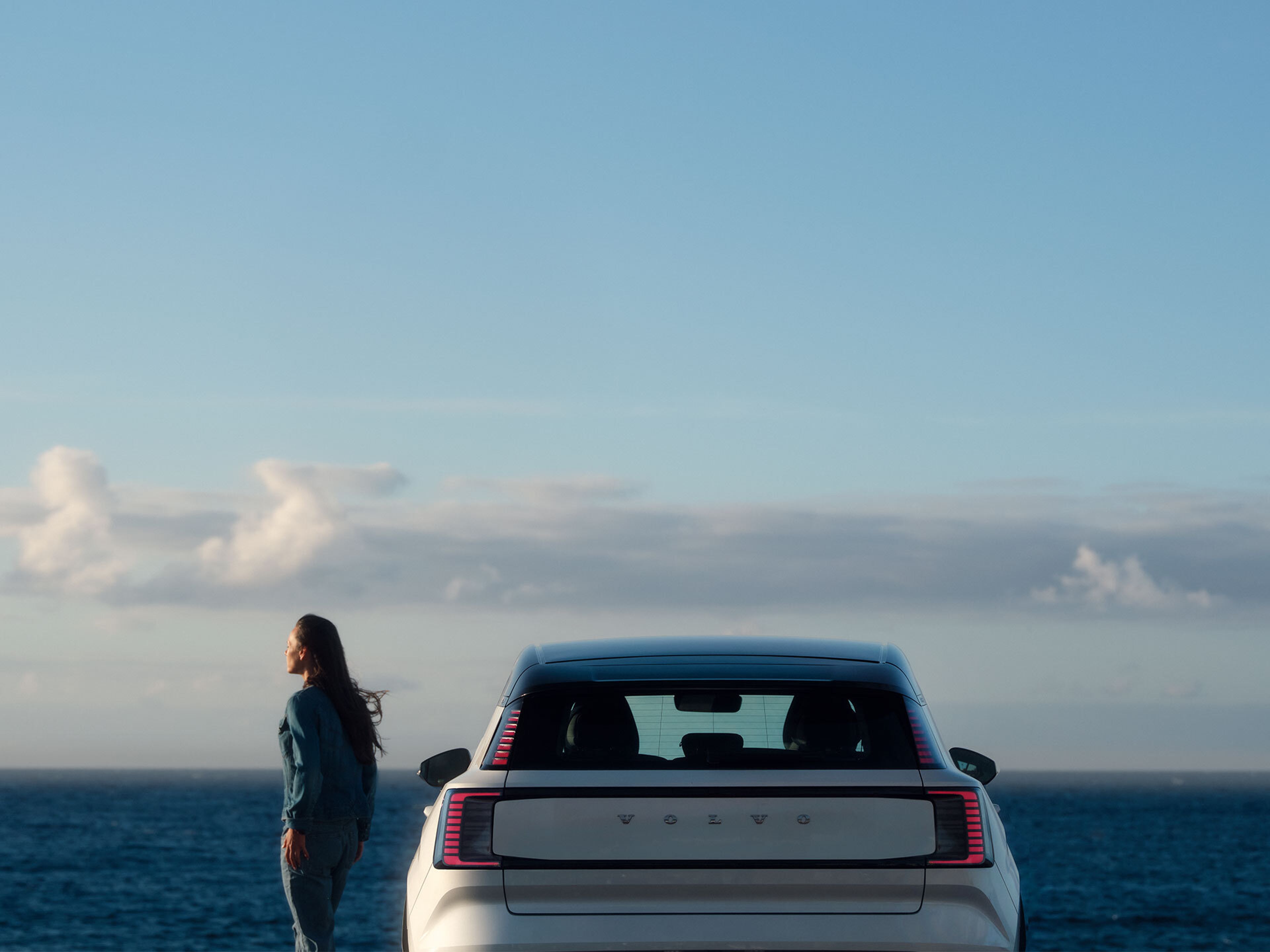 A woman holding open the door of a partially visible Volvo XC90, looking out toward the ocean