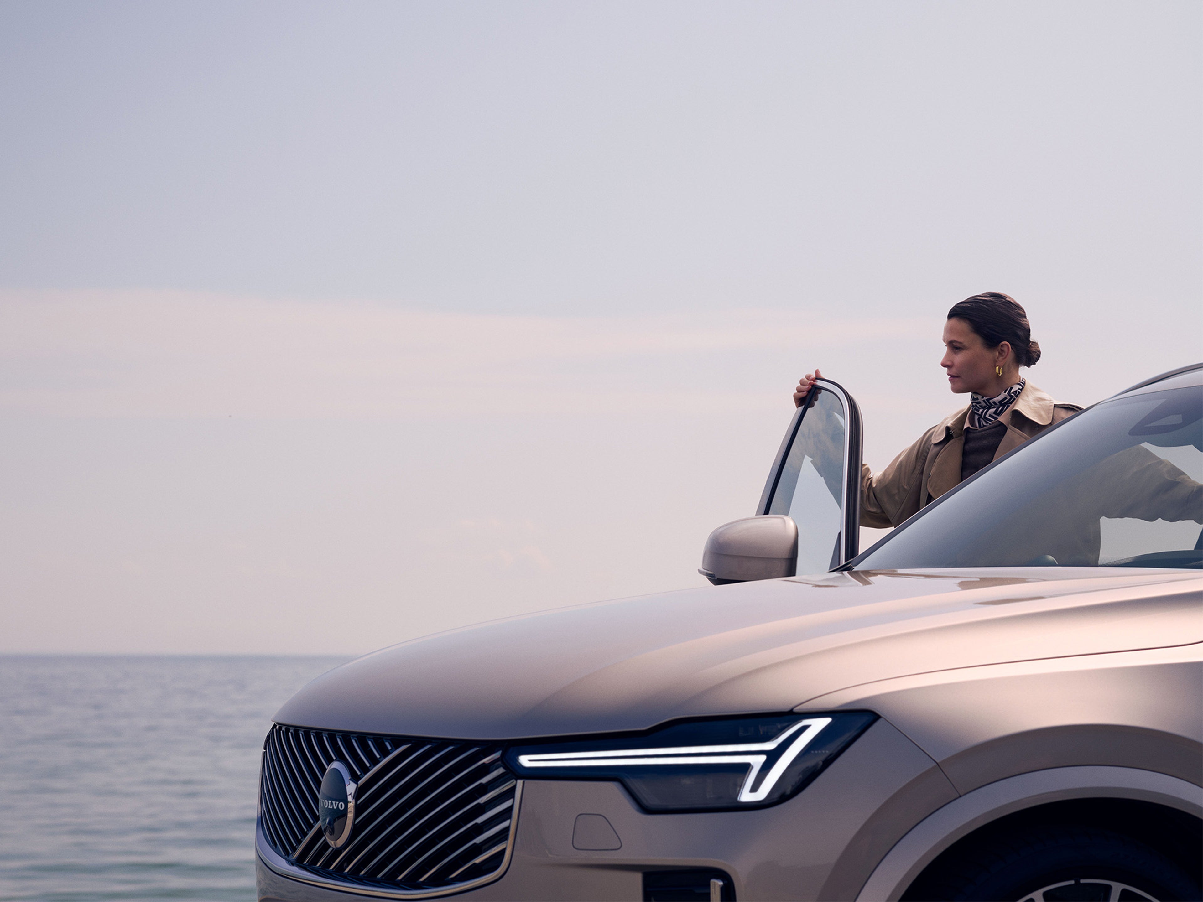 A woman holding open the door of a partially visible Volvo XC90, looking out towards the ocean