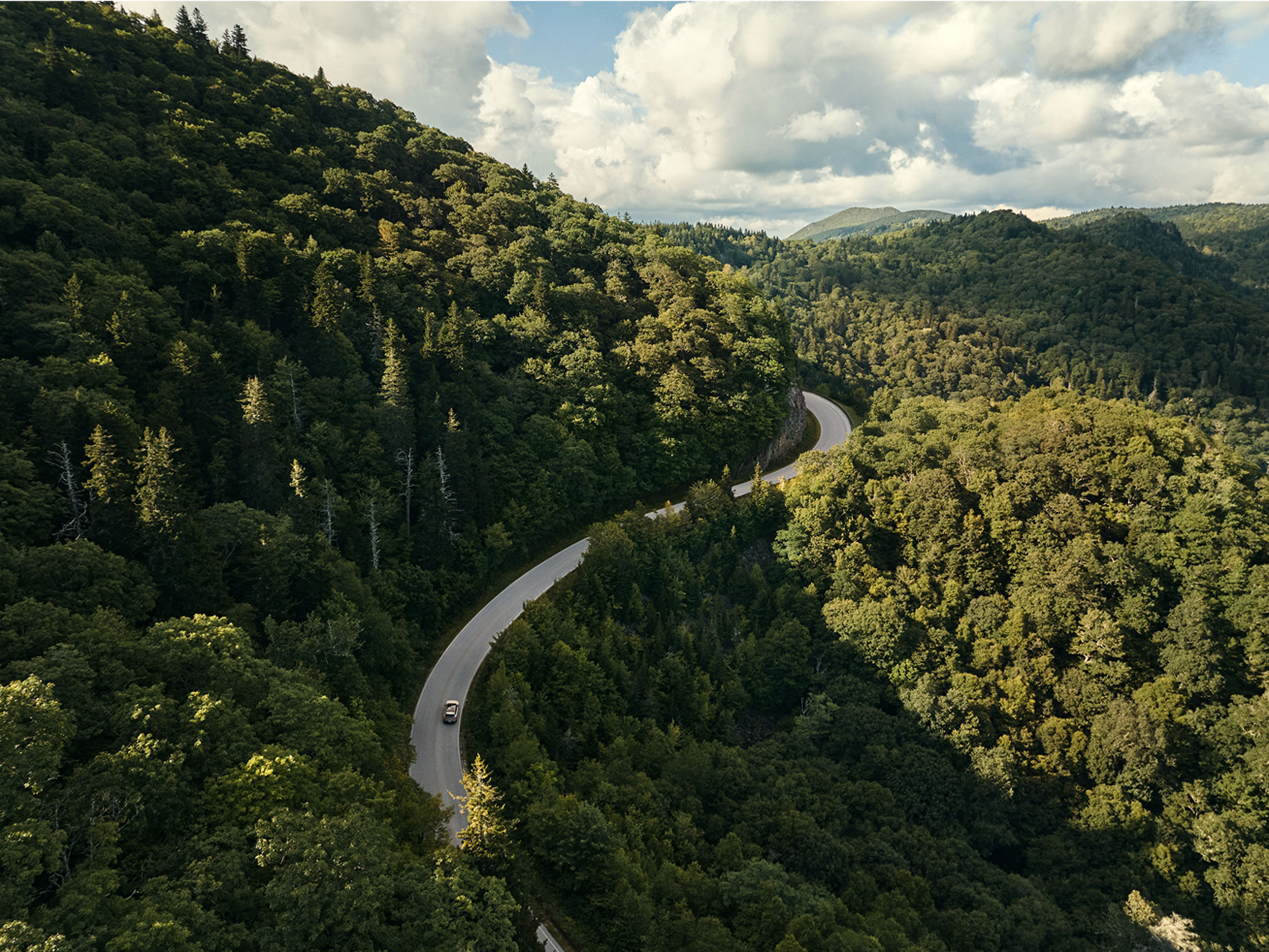 Luchtfoto van een Volvo EX90 die over een bochtige weg rijdt, door dichte, groene beboste heuvels onder een gedeeltelijk bewolkte hemel. De weg slingert sierlijk door het landschap, omgeven door weelderige natuur.