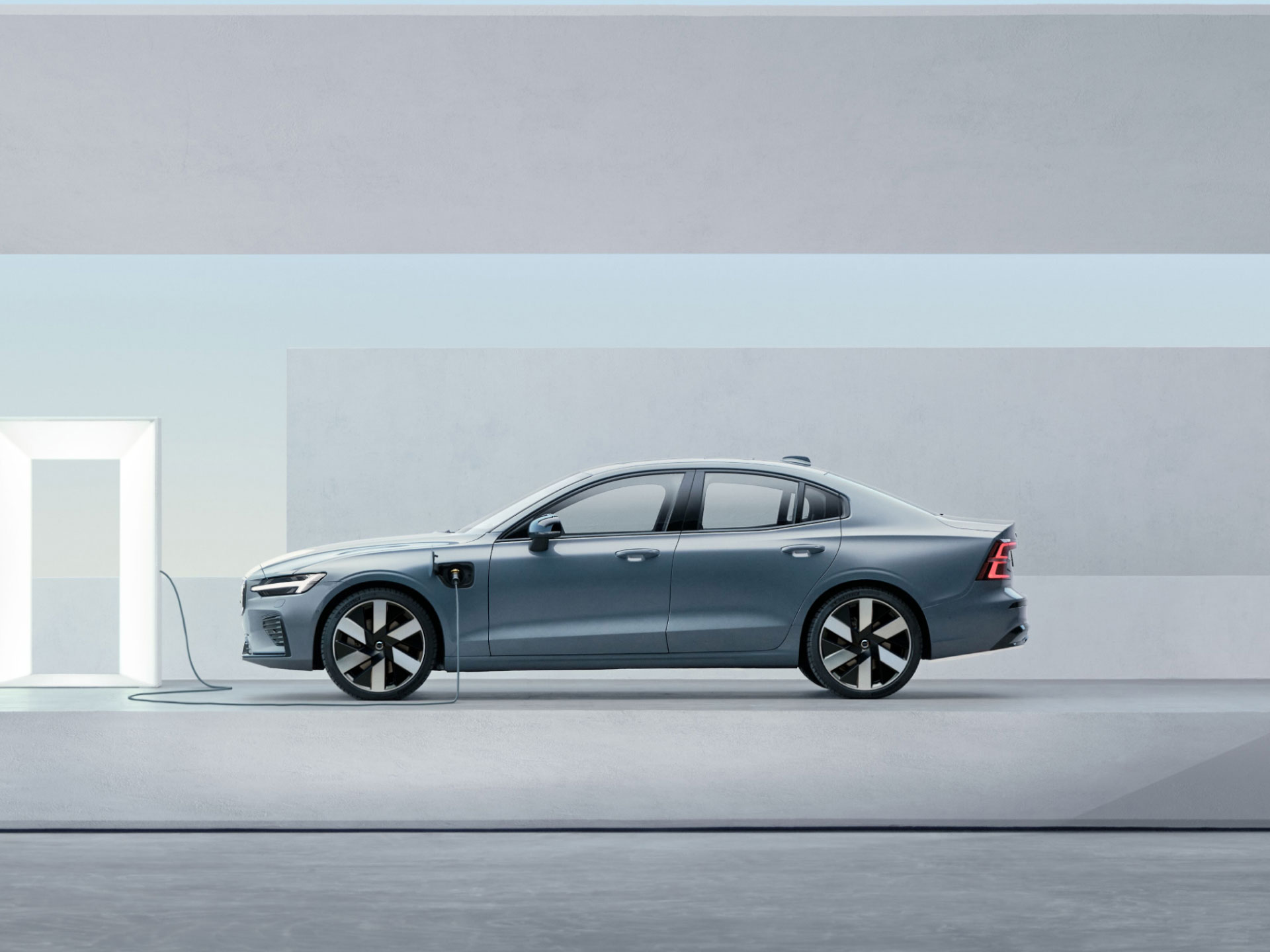 ภาพมุมกว้างด้านข้างของรถยนต์ซีดานระบบปลั๊กอินไฮบริดของ Volvo ขณะเสียบสายอยู่กับเครื่องชาร์จ