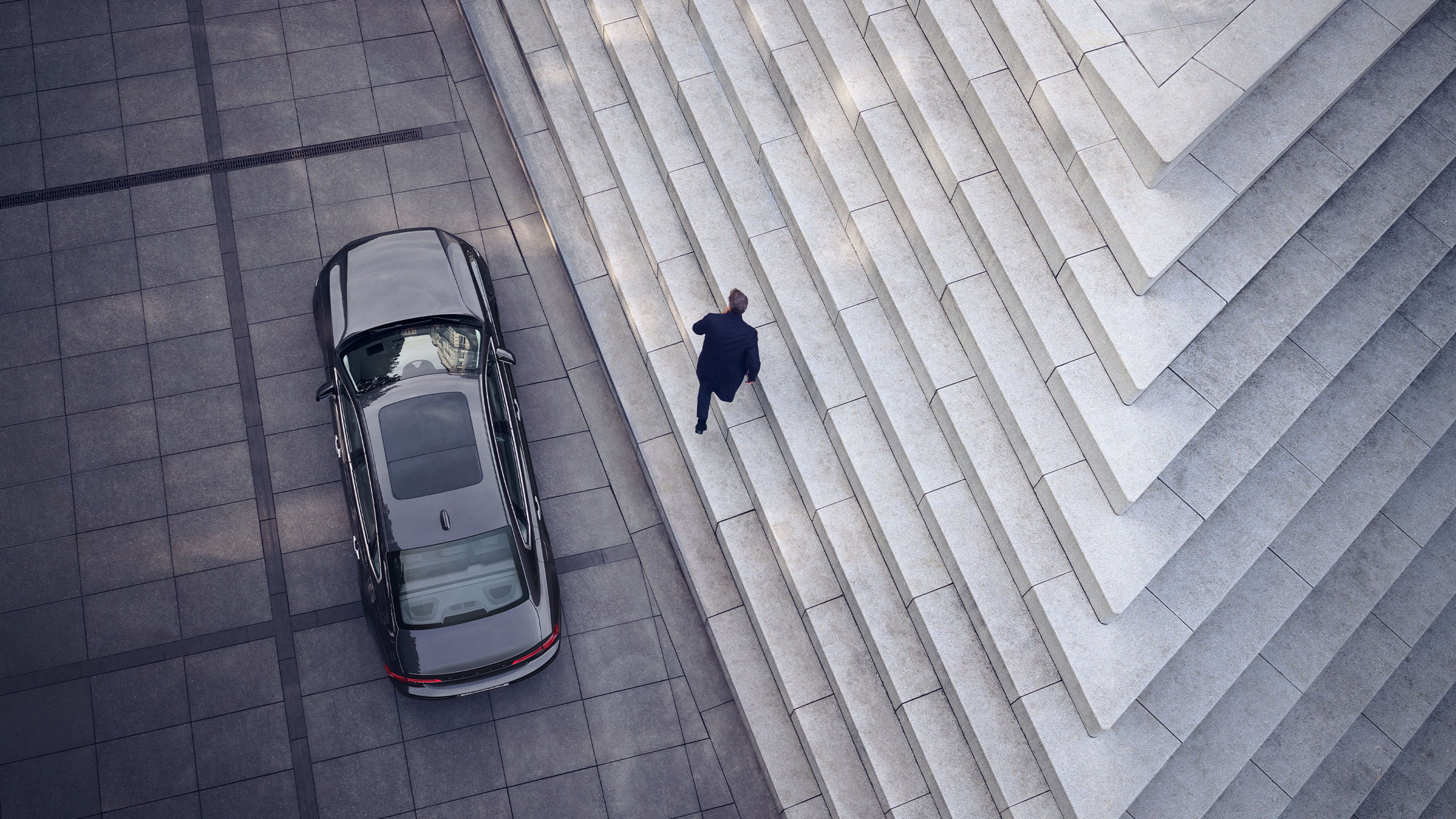 A Volvo S90 sedan is parked beside large, modern stone steps as a person in dark clothing ascends the stairs in an urban setting, viewed from above.