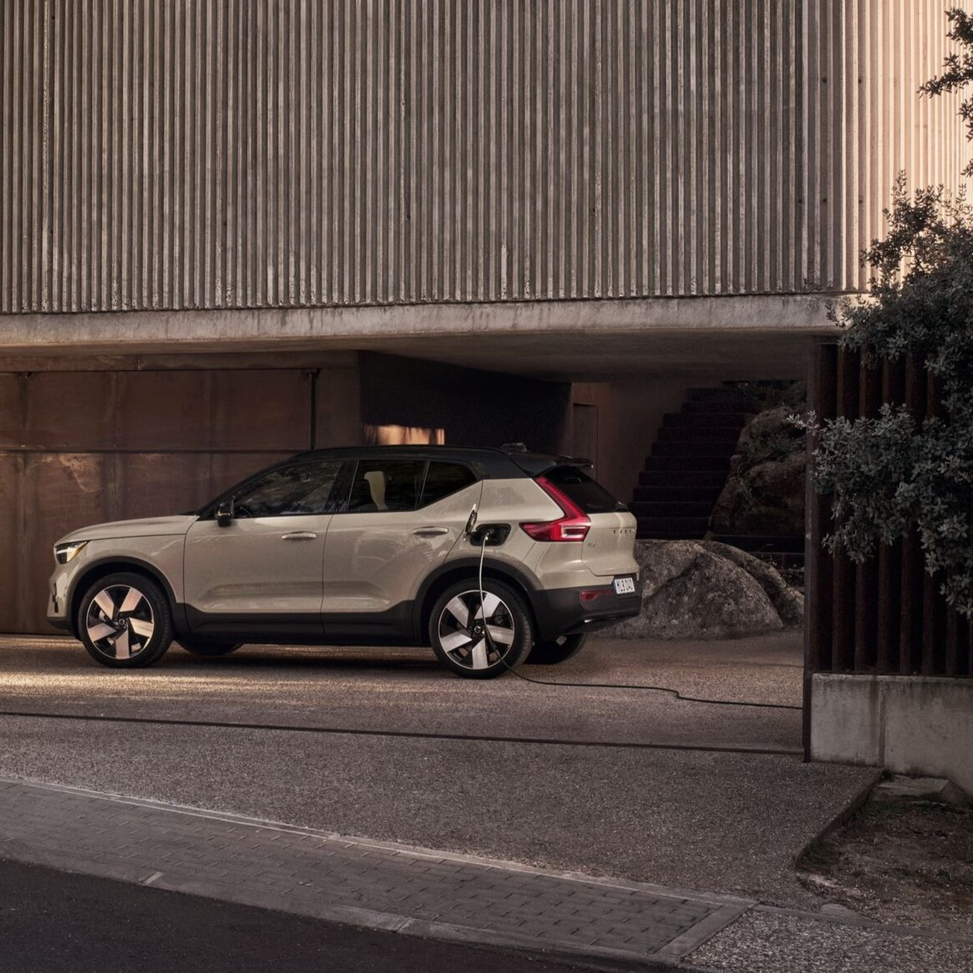 Rear side view of a an electric Volvo SUV charging at a 
home wallbox against a dark wooden wall.