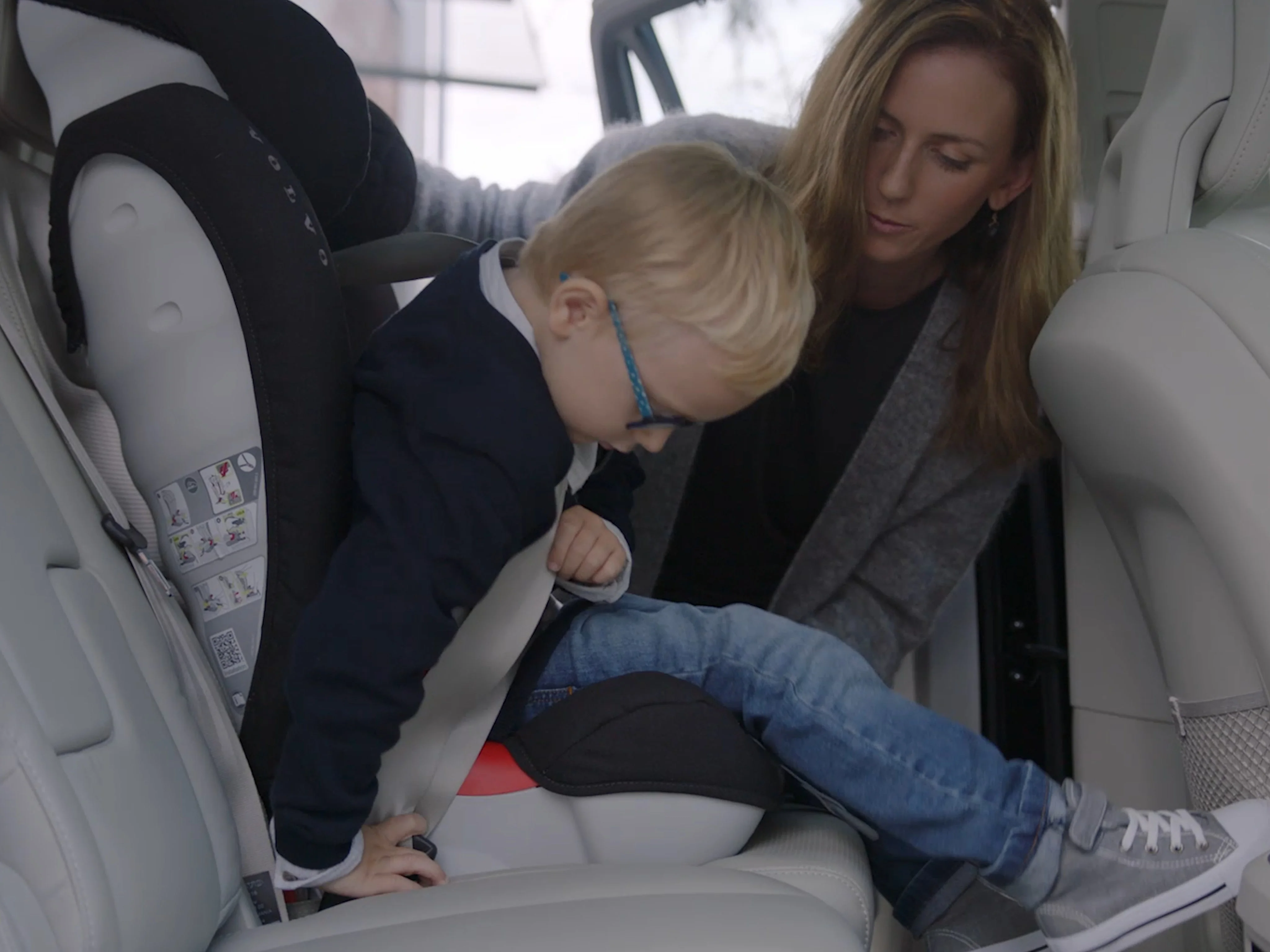 A small child is helped by a woman to buckle into a car seat in the back of a Volvo car.