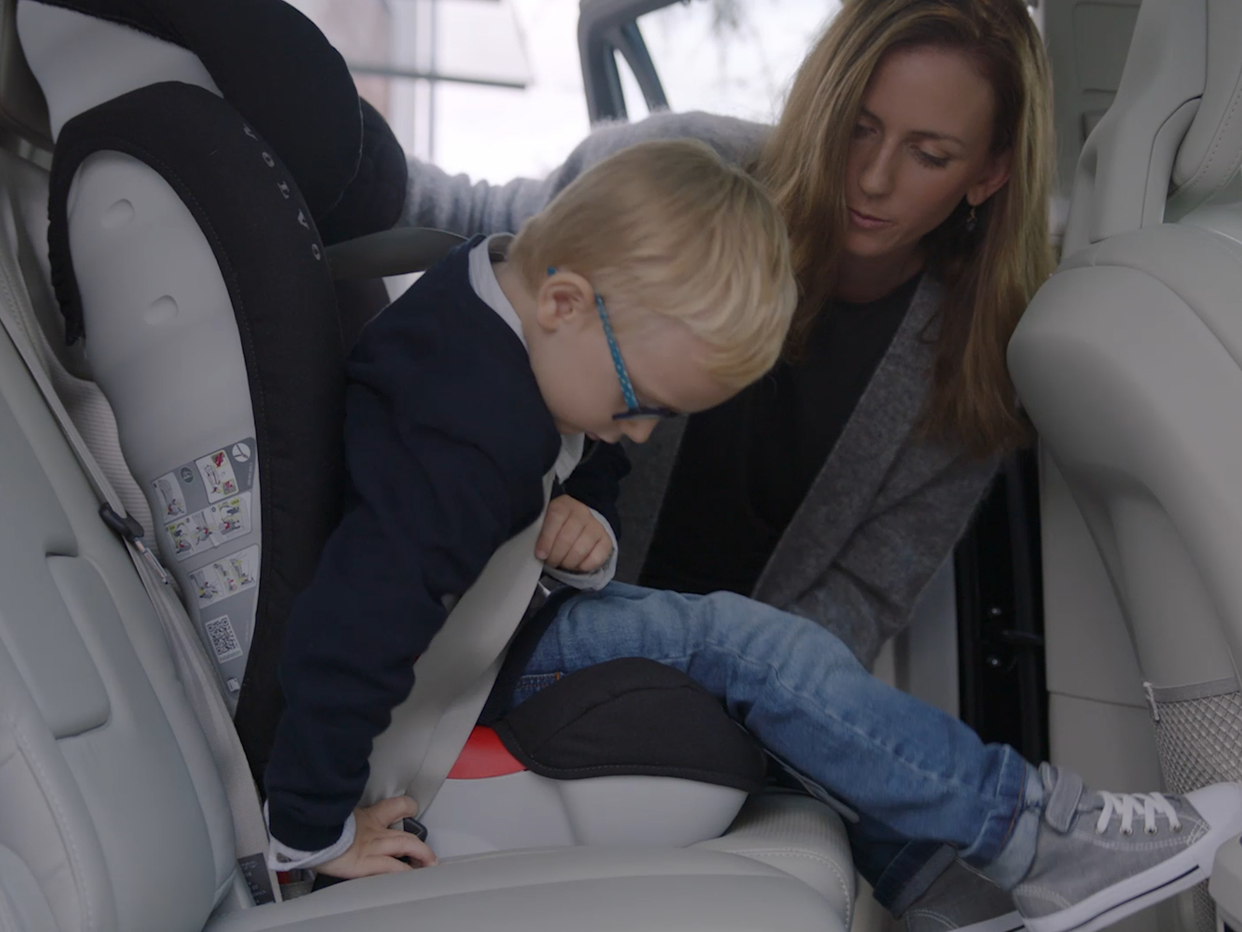 A small child is helped by a woman to buckle into a car seat in the back of a Volvo car. 