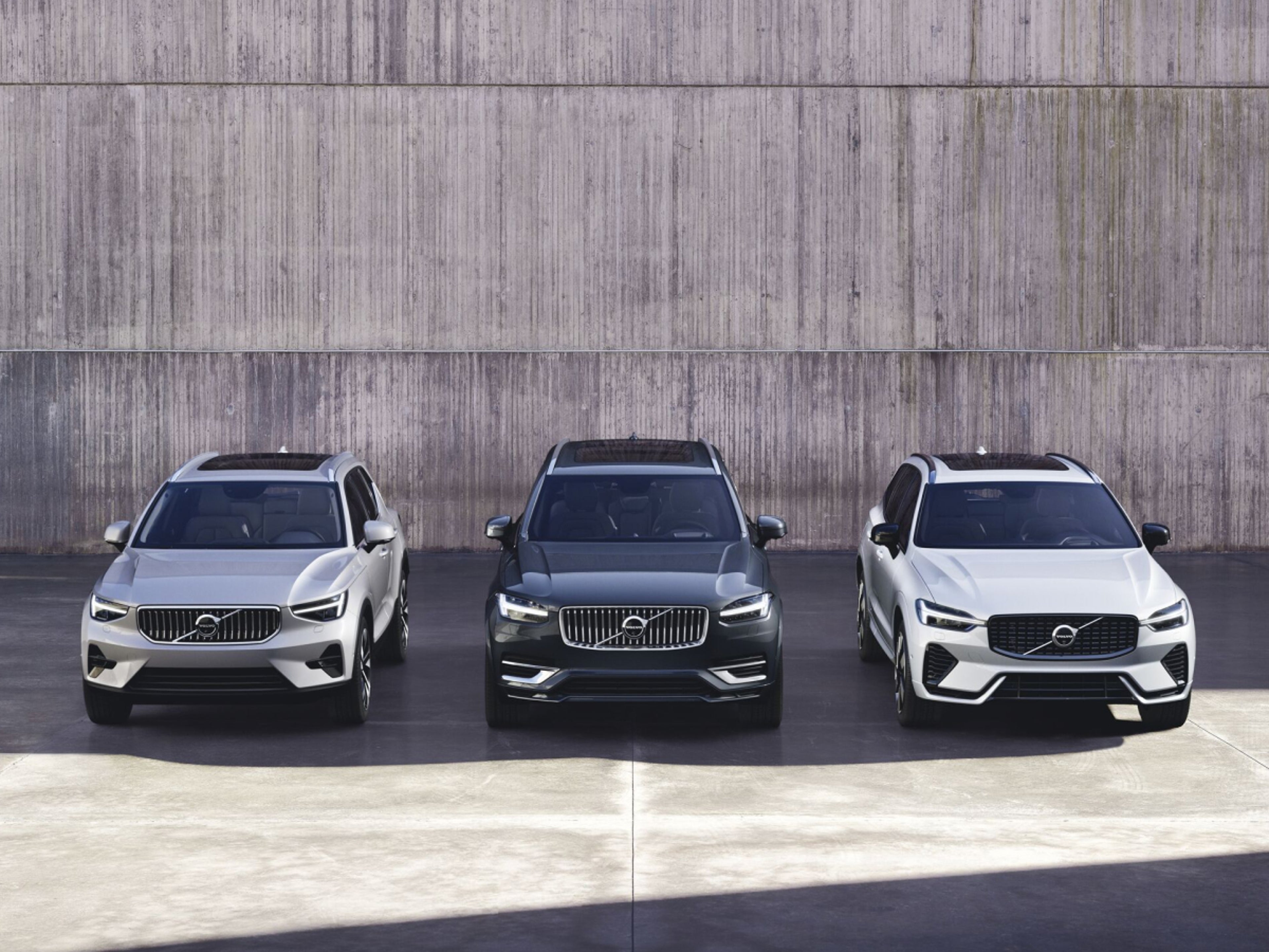 Vue de face de trois SUV Volvo - les XC40, XC90 et XC60 - garés côte à côte devant un mur en béton.