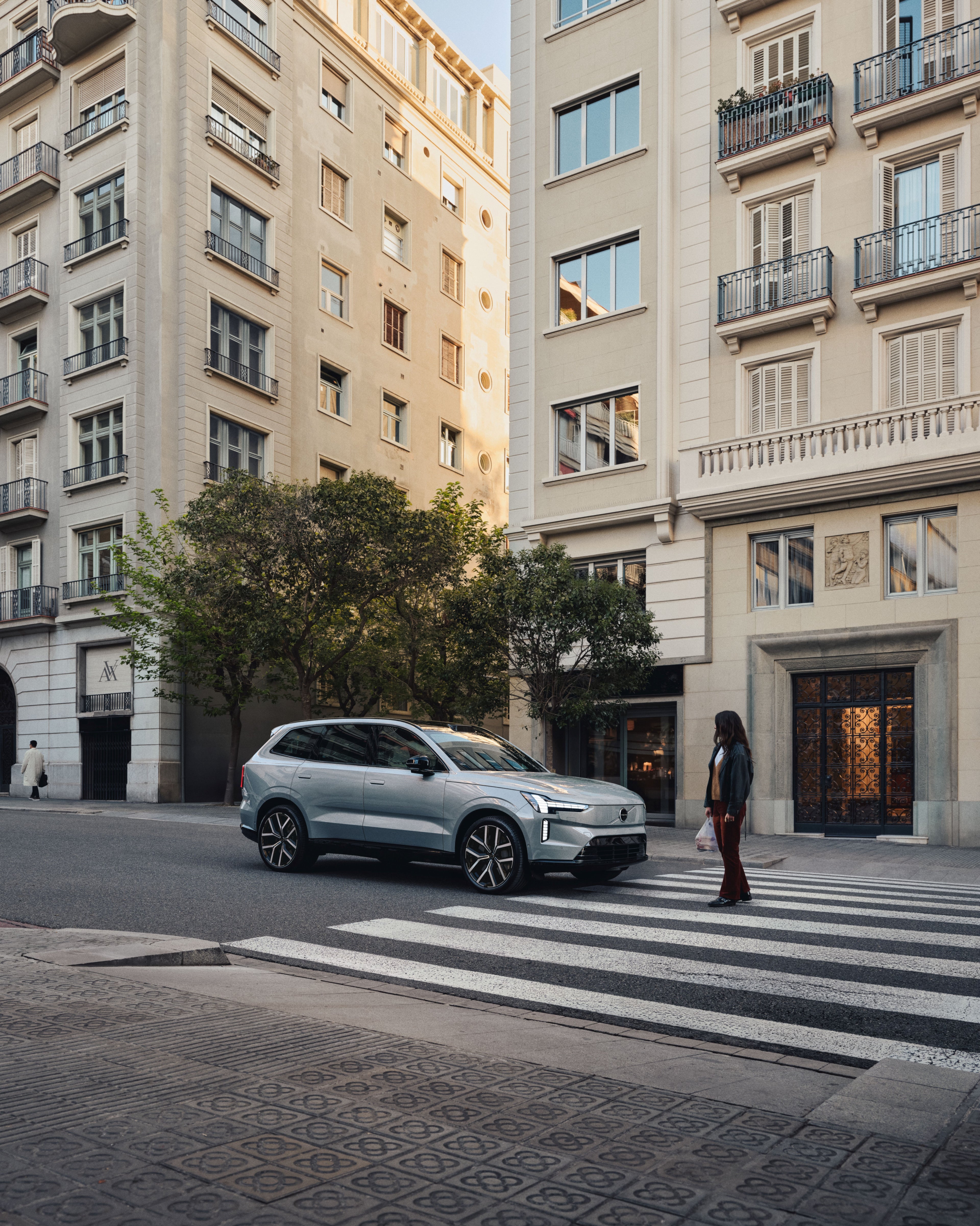 A blurred woman stands in the middle of a street with a Volvo EX90 visible behind her.