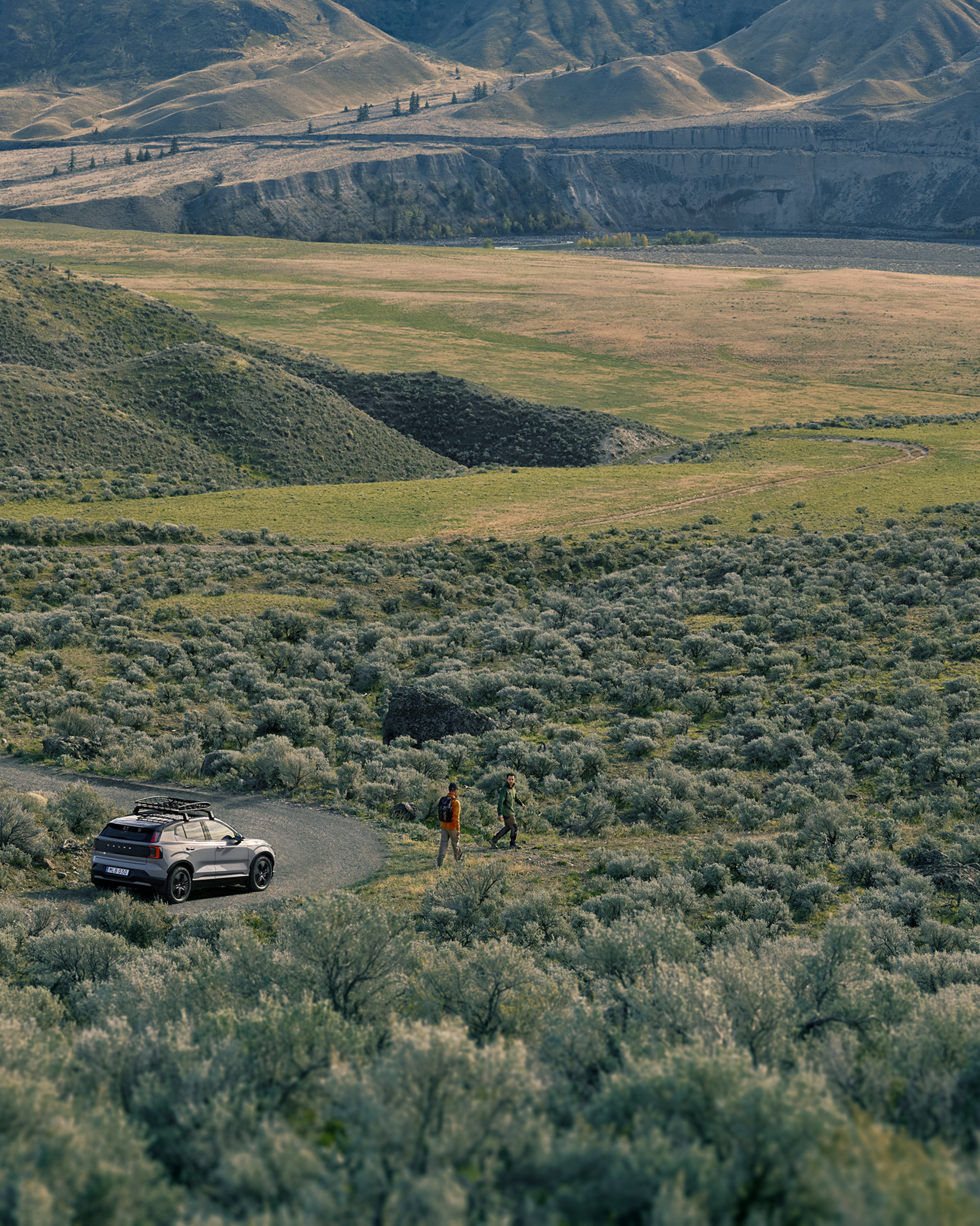 Two people have parked their Volvo EX30 on a gravel road and are walking into a vast, scenic landscape of rolling hills and open nature.