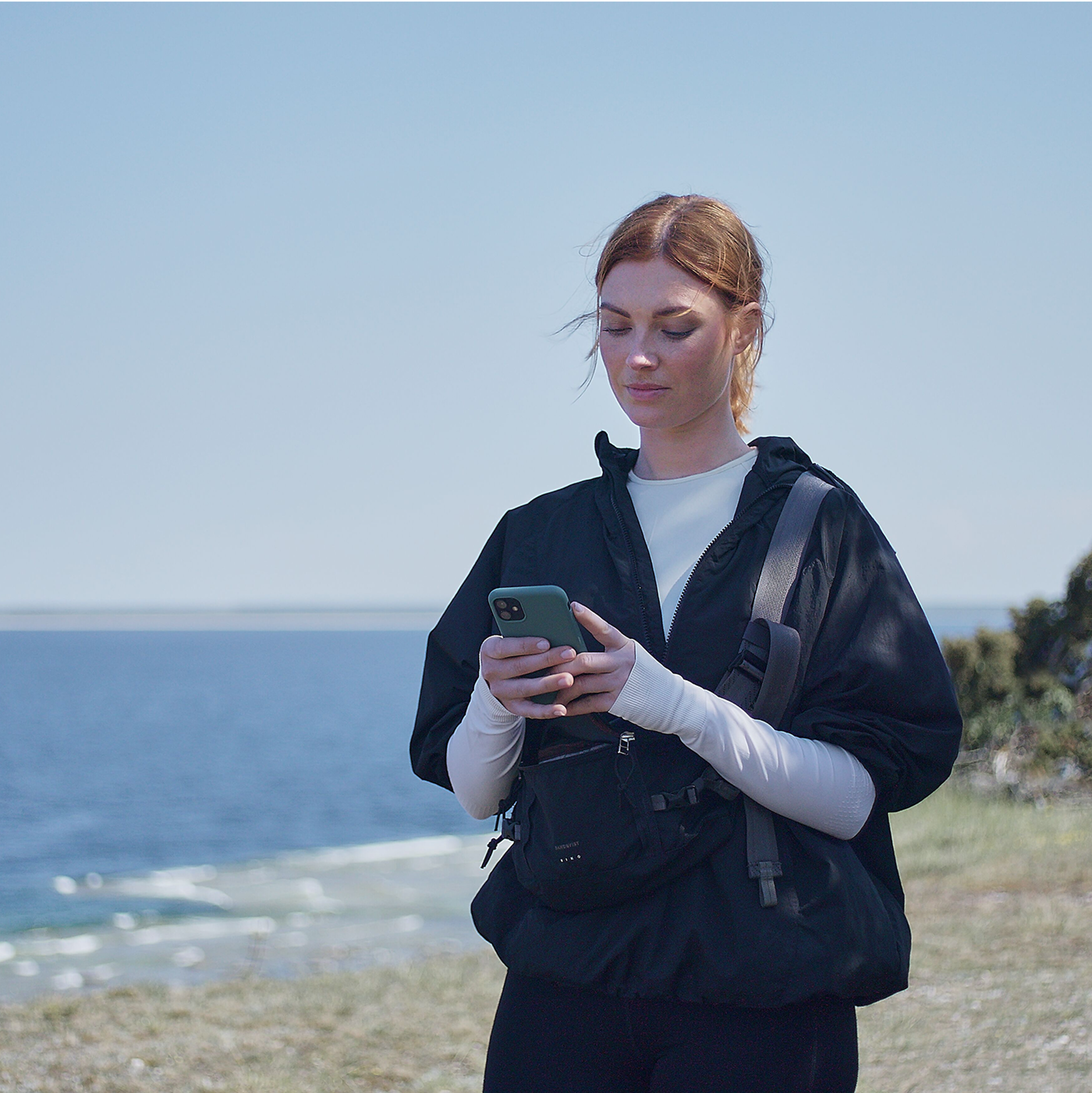 Vrouw die bij de zee staat en haar smartphone gebruikt terwijl ze een zwarte jas en een crossbodytas draagt.