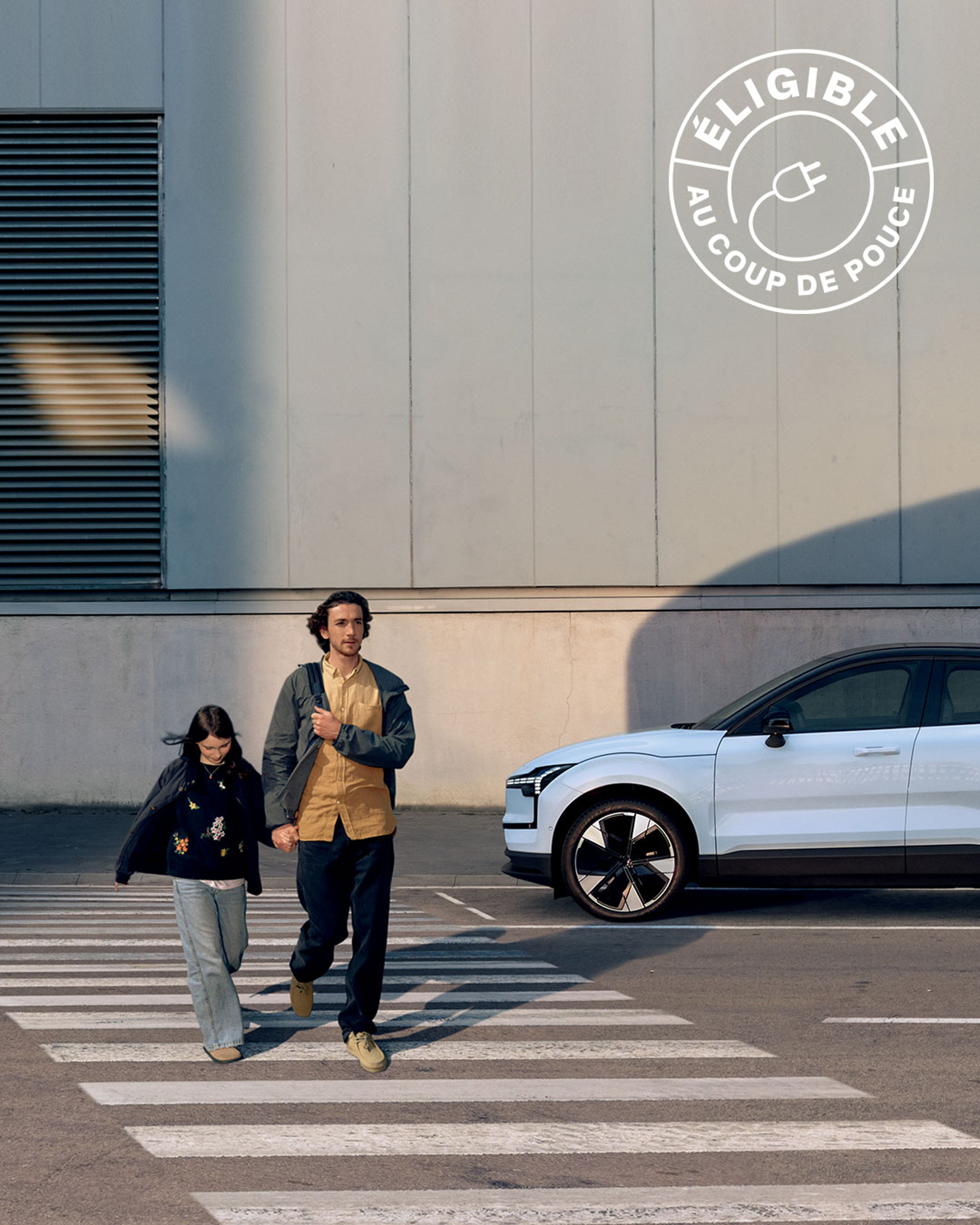 A white Volvo EX30 electric SUV is parked and charging at a station outdoors, with a person in a light coat standing in the foreground, partially visible from behind.