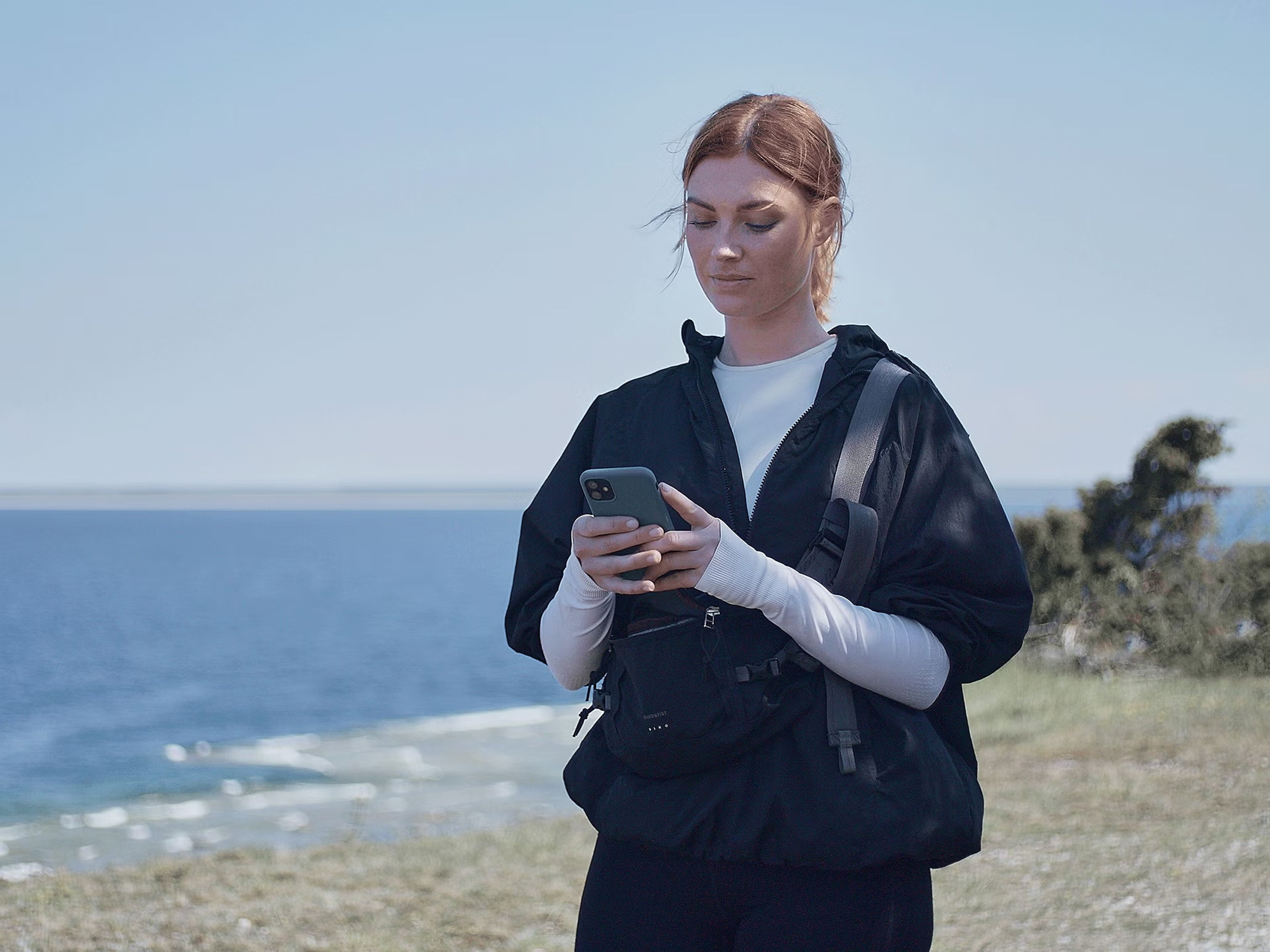 Eine rothaarige Frau steht an einer Küste und schaut auf Ihr Handy. Sie liest gespannt etwas darauf.