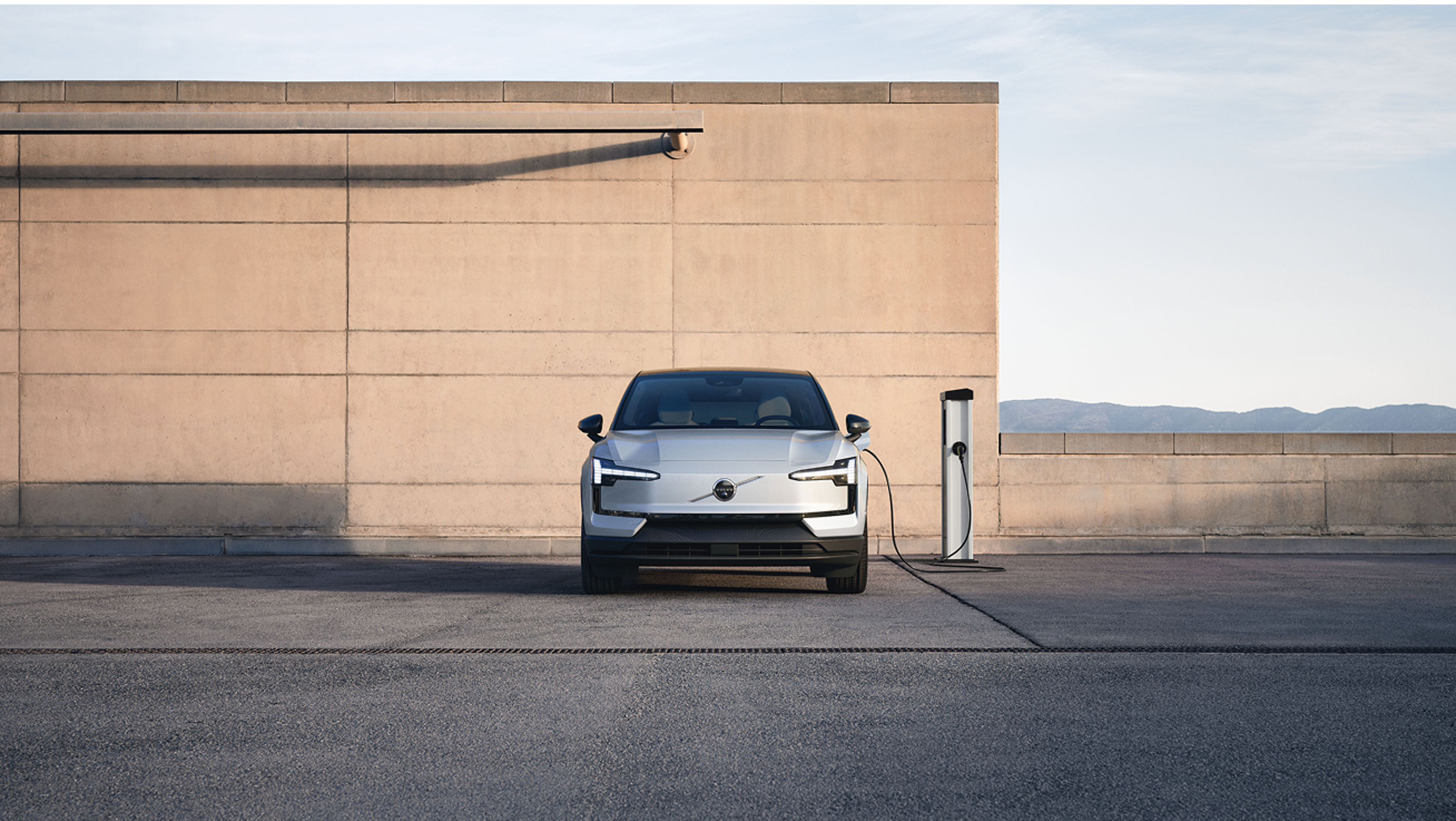 Skats no priekšpuses uz elektrisko Volvo automašīnu, kas saulainā dienā pievienota publiskajam uzlādes punktam autostāvvietā.