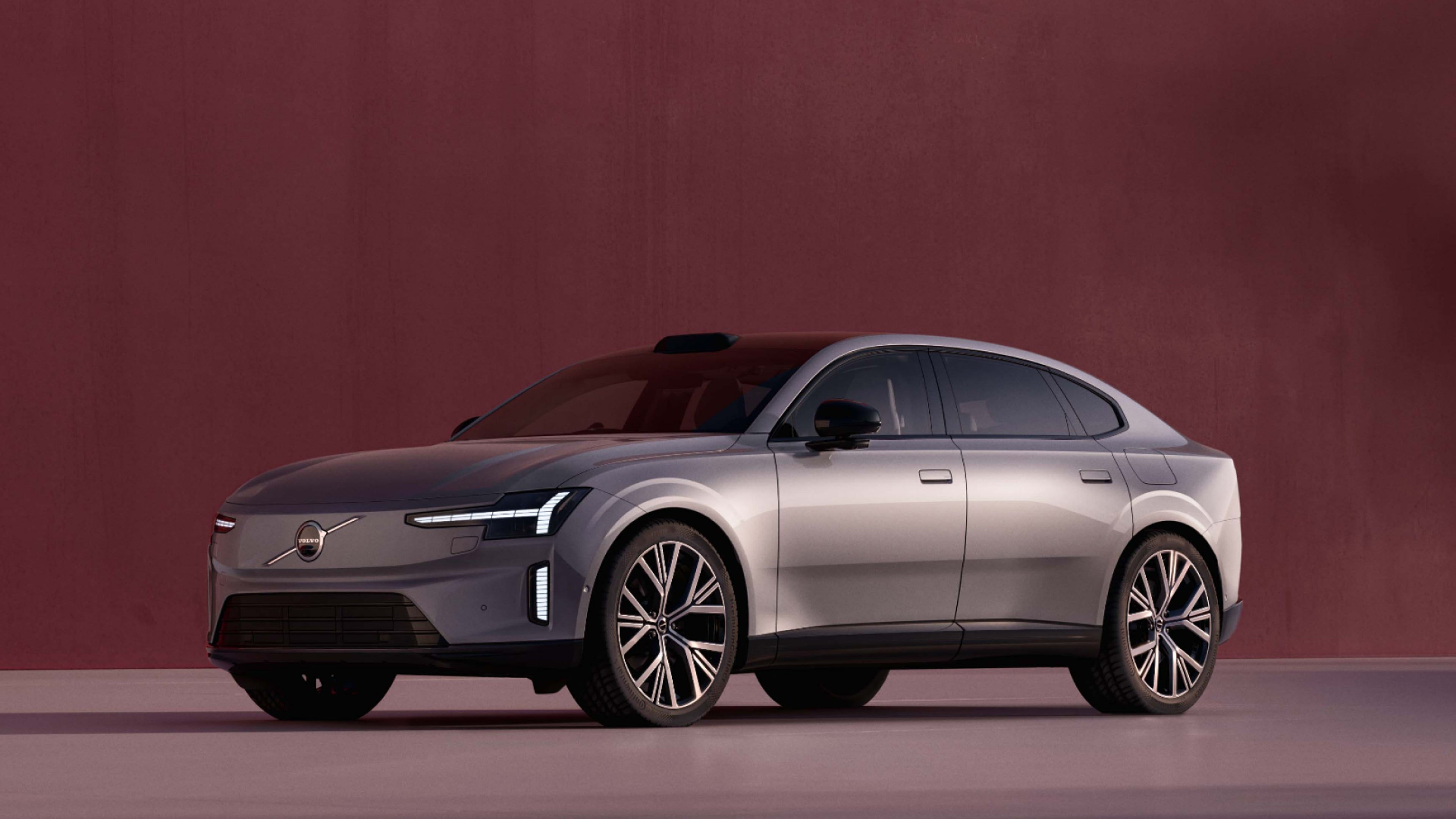 Egy szürke Volvo ES90 oldalnézetből egy nyitott, üres belső térben, sötét vörös háttérrel.