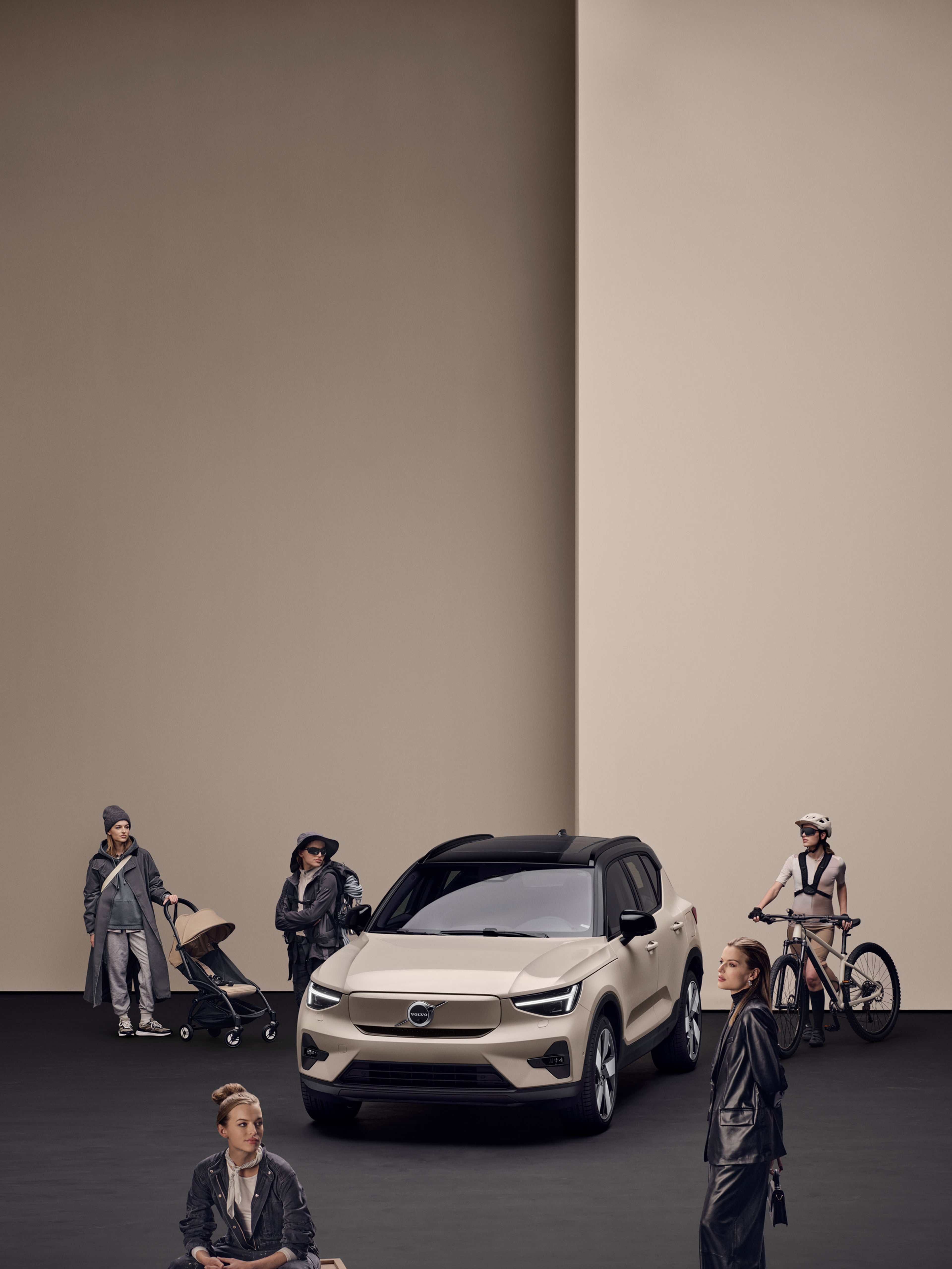 Beige Volvo EX40 elbil i en studio med svart golv och beige bakgrund, omgiven av personer i olika livsstilssituationer, inklusive en cyklist, en person med barnvagn och andra i vardagskläder.