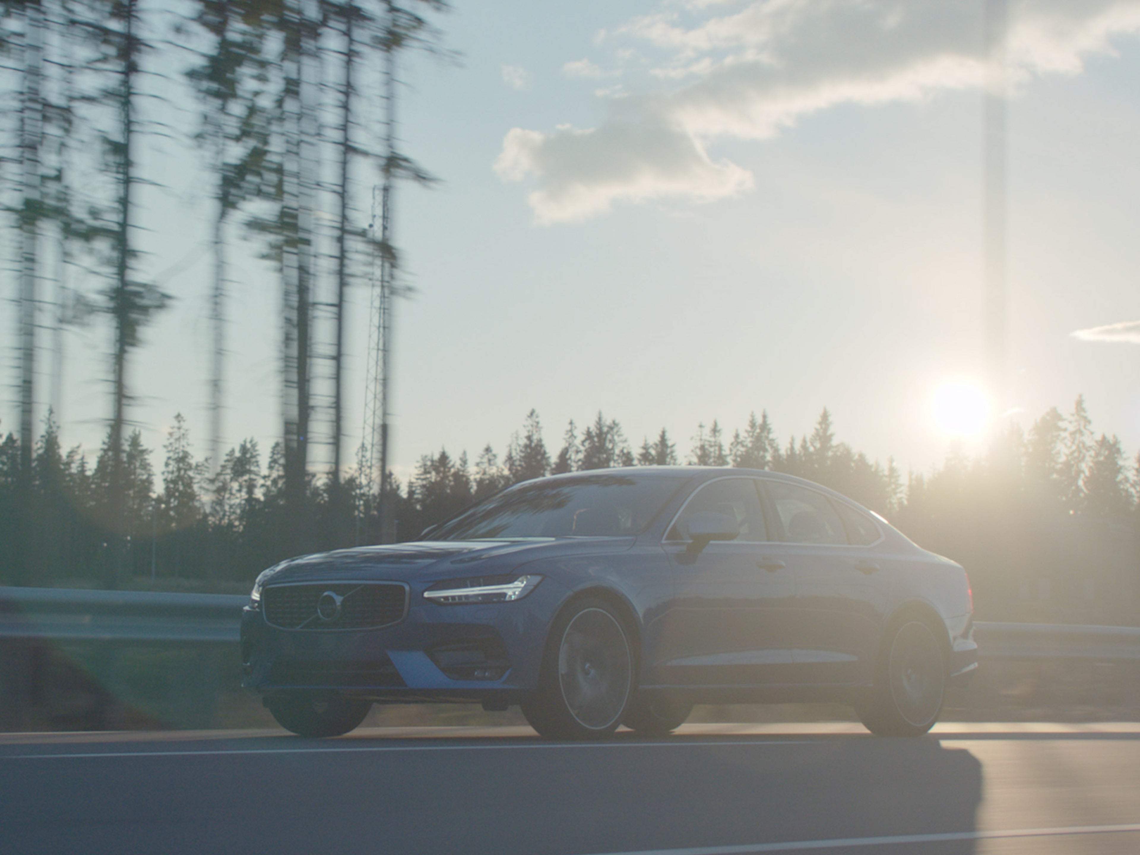 Platus saulėtą dieną miško supamu keliu važiuojančio „Denim Blue“ spalvos „Volvo S90“ sedano kadras.