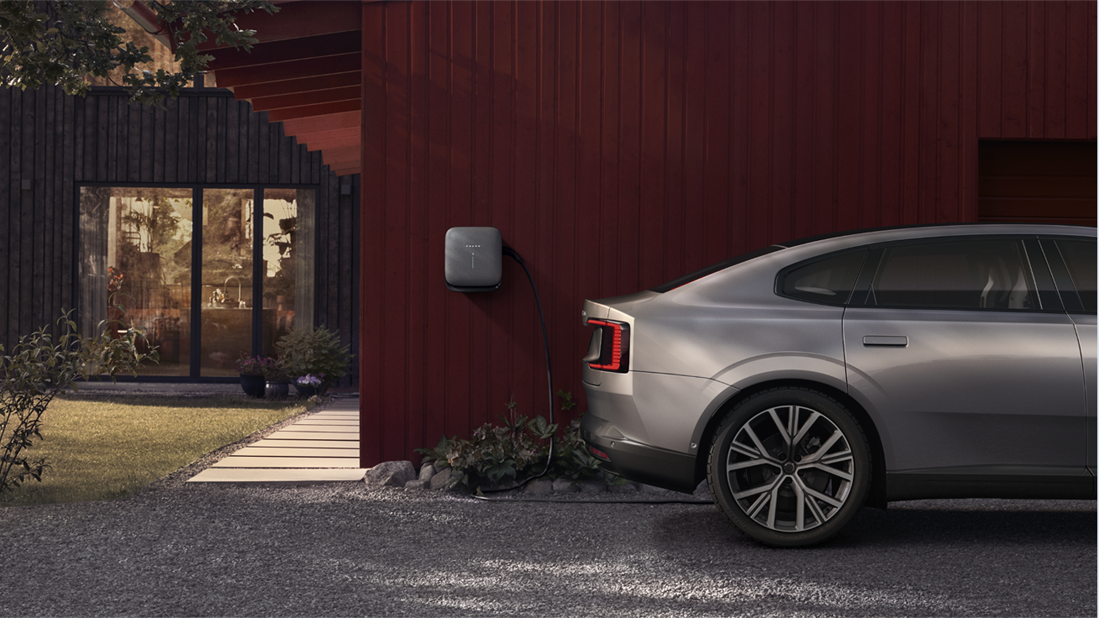 Sānskats uz elektrisko Volvo automašīnu, kas pievienota mājas
sienas uzlādes punktam pret sarkanu mājas ārpusi.