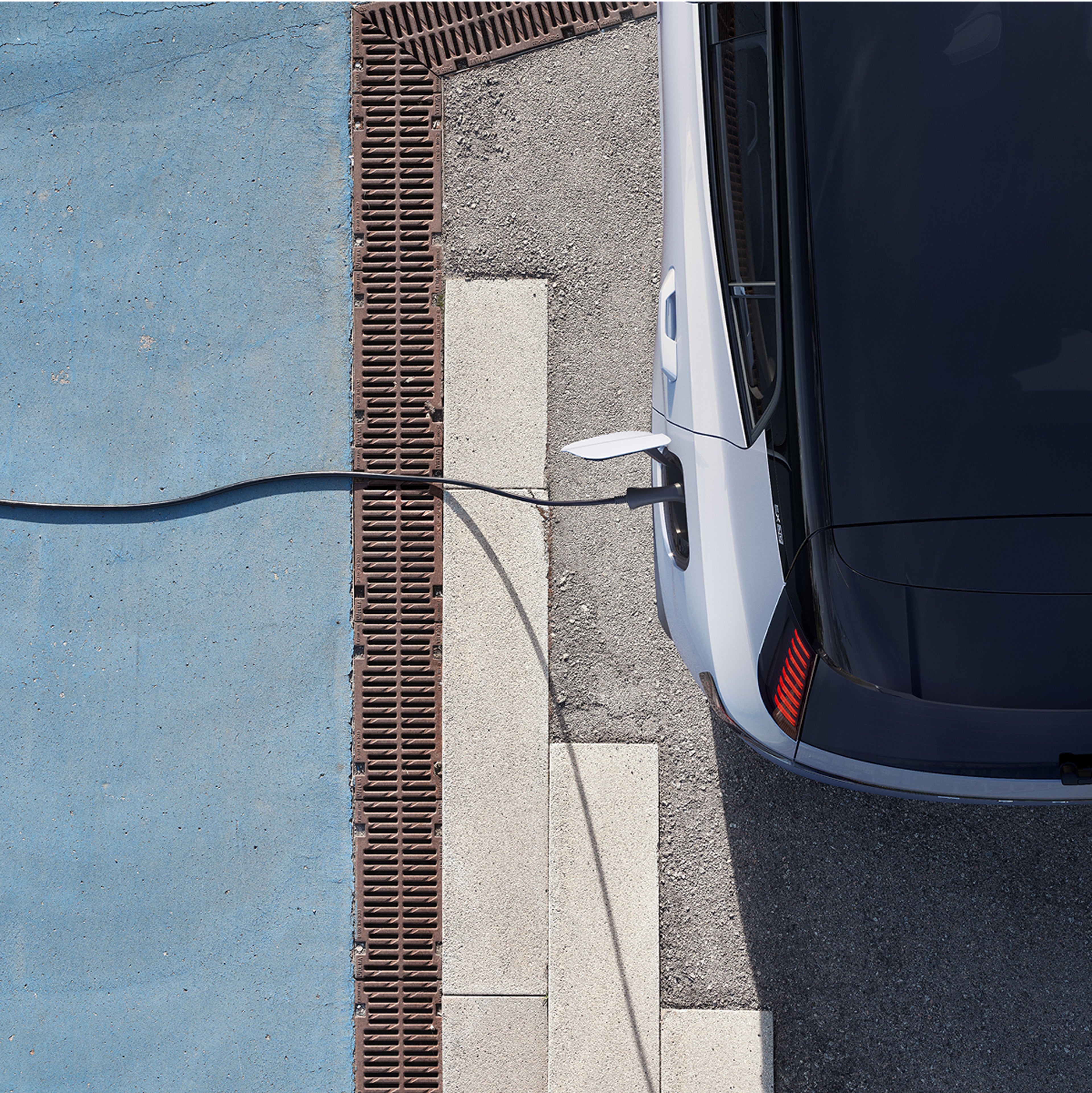 Top birds eye view of an electric SUV with a charging cable 
plugged into its left side, running off to the left of the pavement.