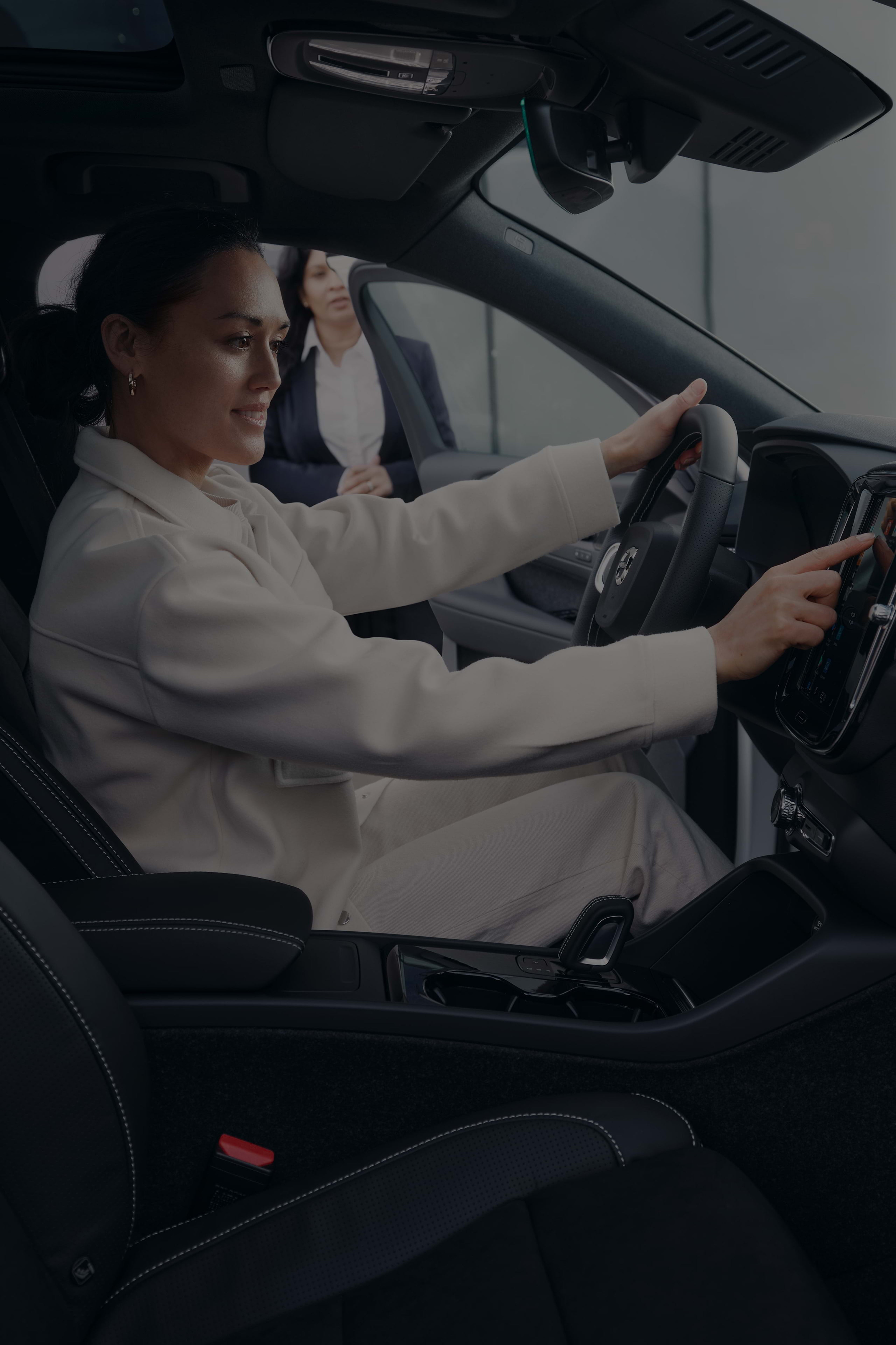 Pardavėjos prižiūrima jauna moteris šypsodamasi sėdi vairuotojo vietoje ir liečia „Volvo“ automobilio centrinę konsolę.