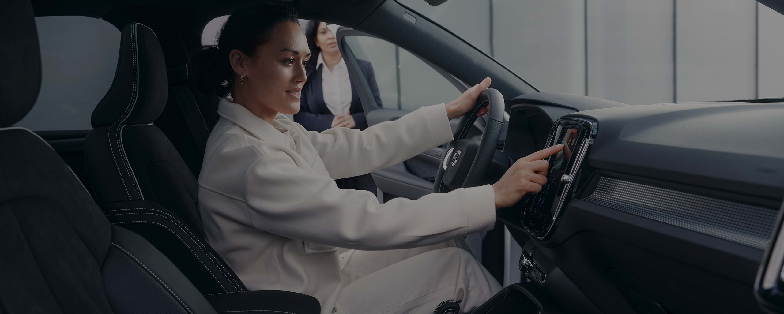 Pardavėjos prižiūrima jauna moteris šypsodamasi sėdi vairuotojo vietoje ir liečia „Volvo“ automobilio centrinę konsolę.