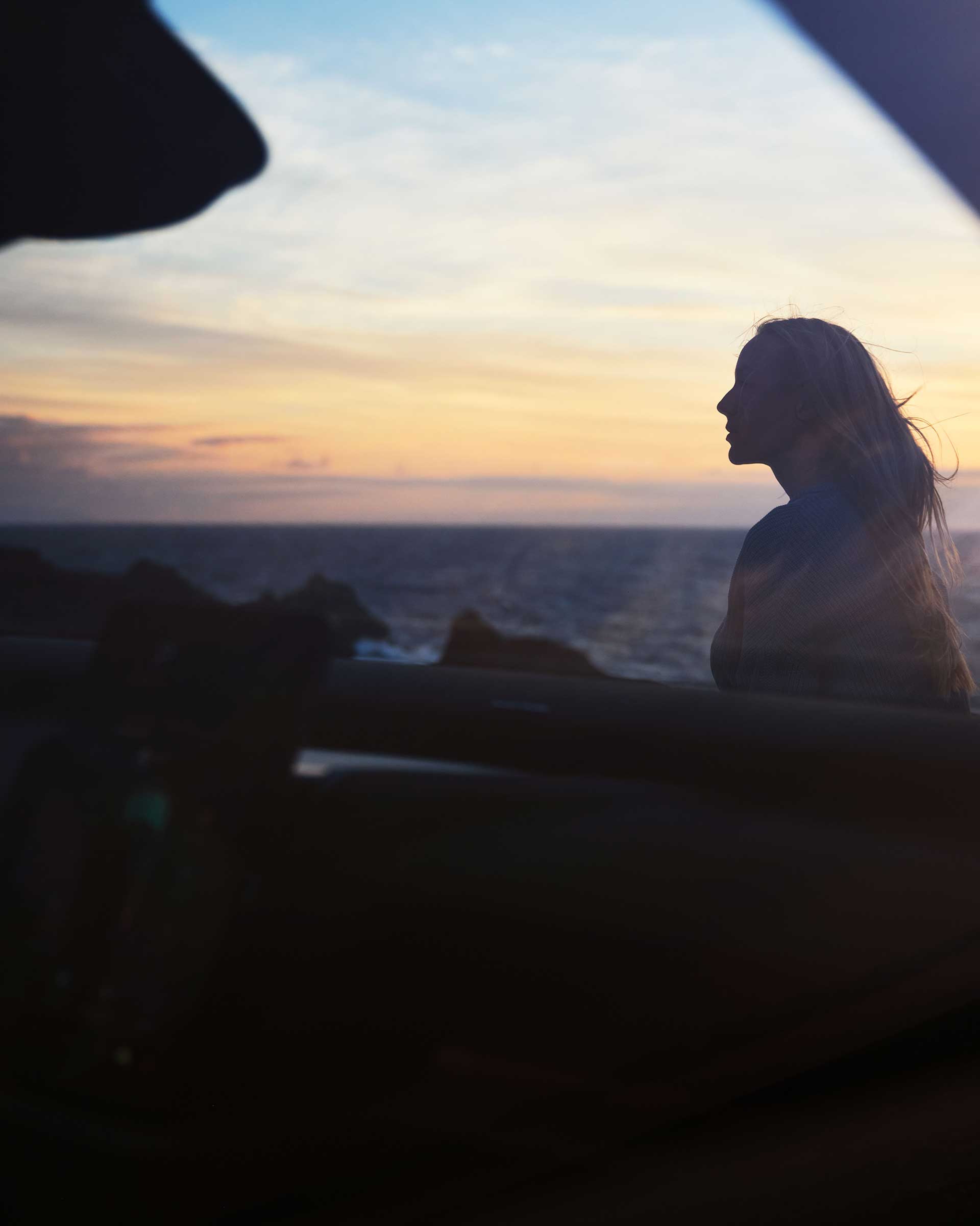 Silhuet af en kvinde med langt hår, der ser på solnedgangen over havet gennem et bilvindue. Stemningen er rolig og eftertænksom.
