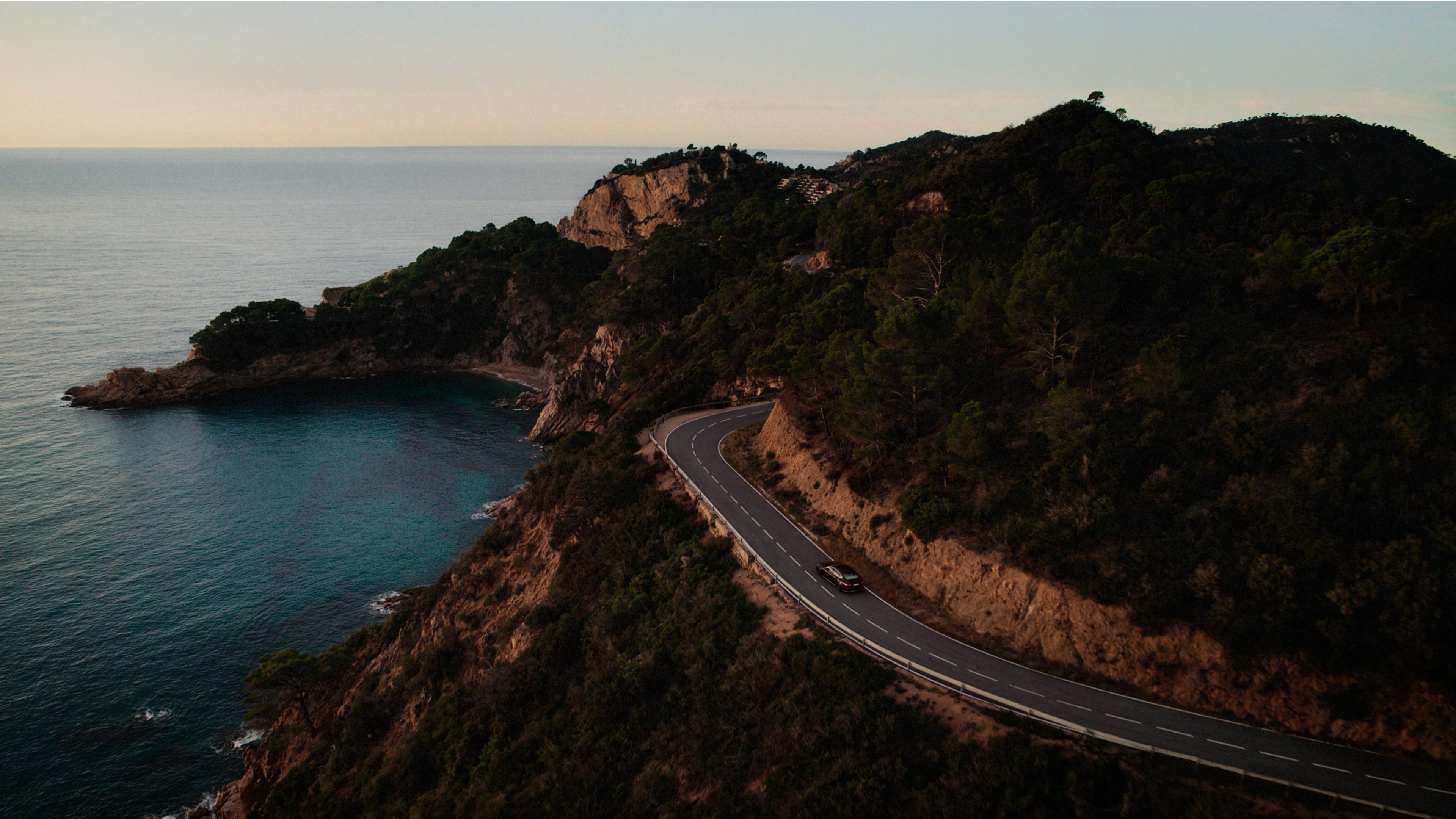 Een auto rijdt over een kronkelende kustweg omringd door kliffen, bomen en turquoise water onder een heldere hemel.