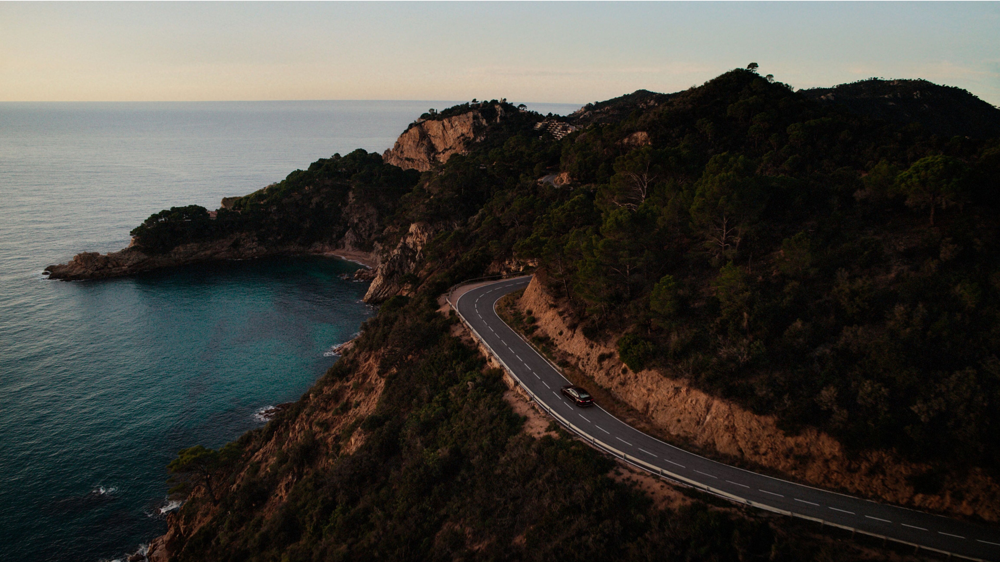 A car drives along a winding coastal road surrounded by cliffs, trees, and turquoise water under a clear sky.