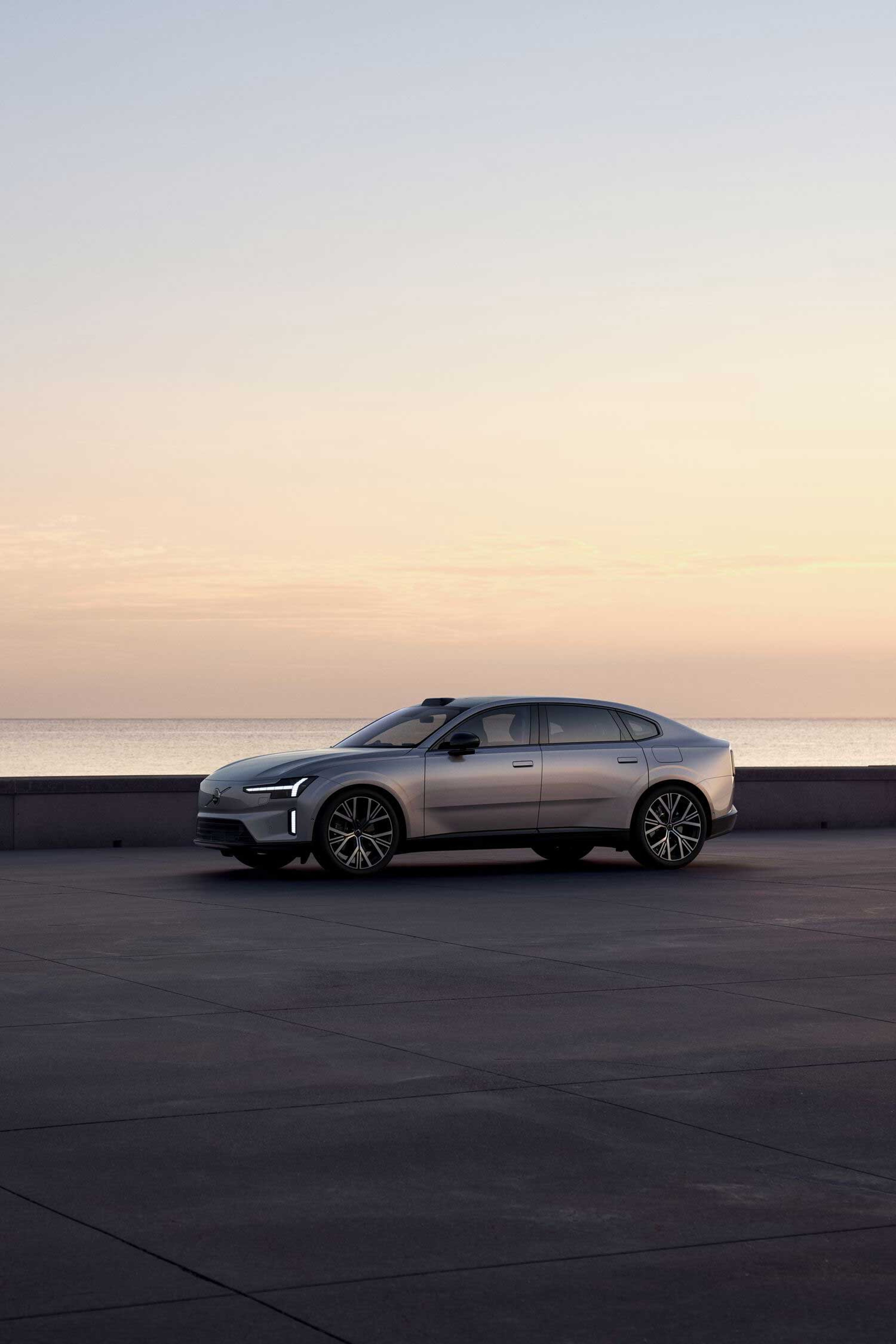 Elegant sølvfarvet Volvo sedan på en stor, tom betonflade nær havet. Den rolige solnedgangshimmel kaster varme nuancer og skaber en fredfyldt og moderne stemning.