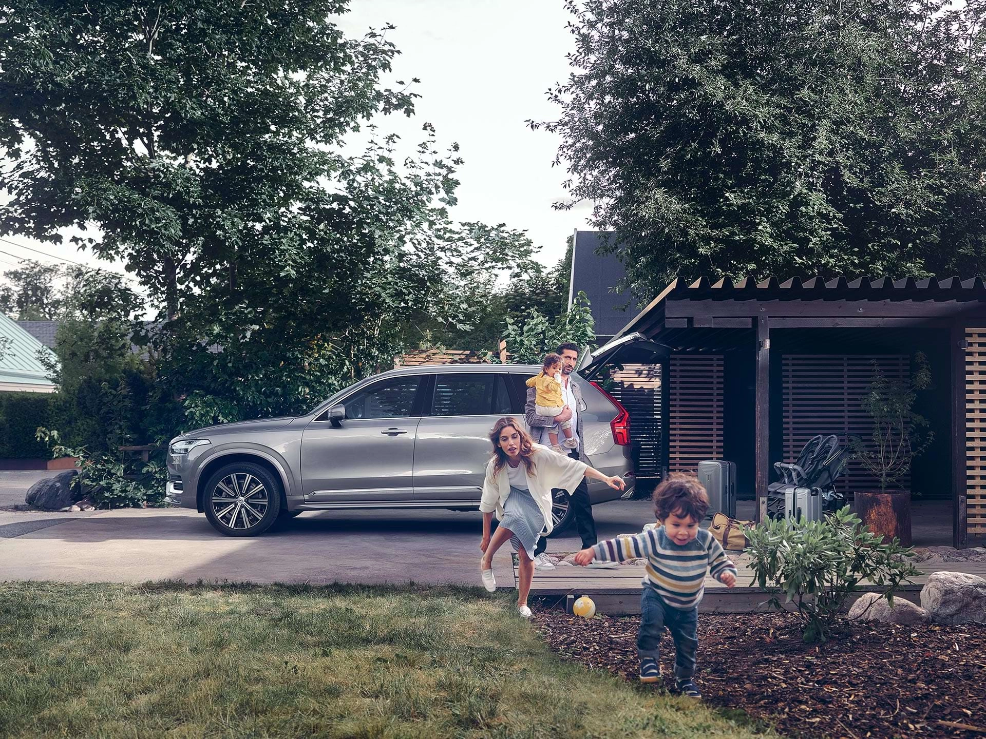 Terwijl een gezin uit een buitenwijk zich voorbereidt op een rit met hun Volvo SUV, rent een kind door de tuin, achtervolgd door zijn moeder.