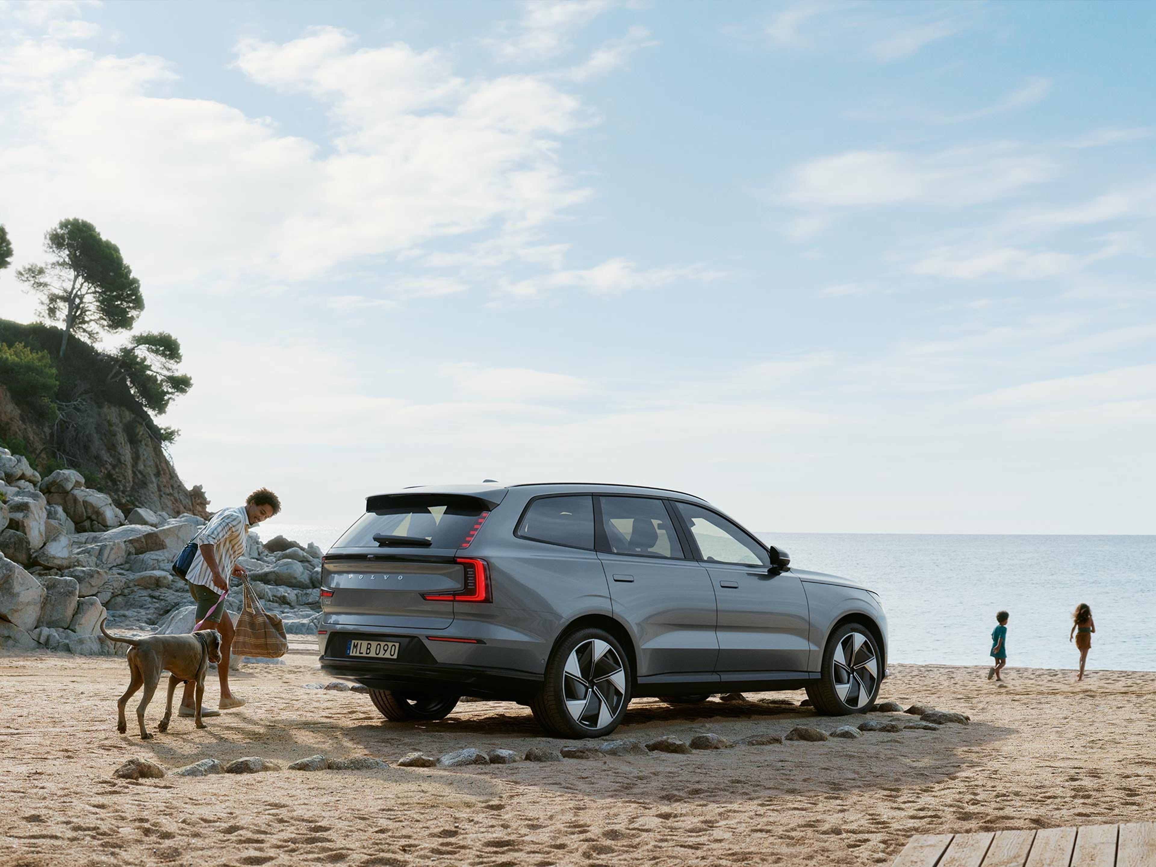 Një burrë drejton qenin e tij të madh drejt detit, ku luajnë fëmijët; Volvo SUV e familjes qëndron e parkuar në rërë.