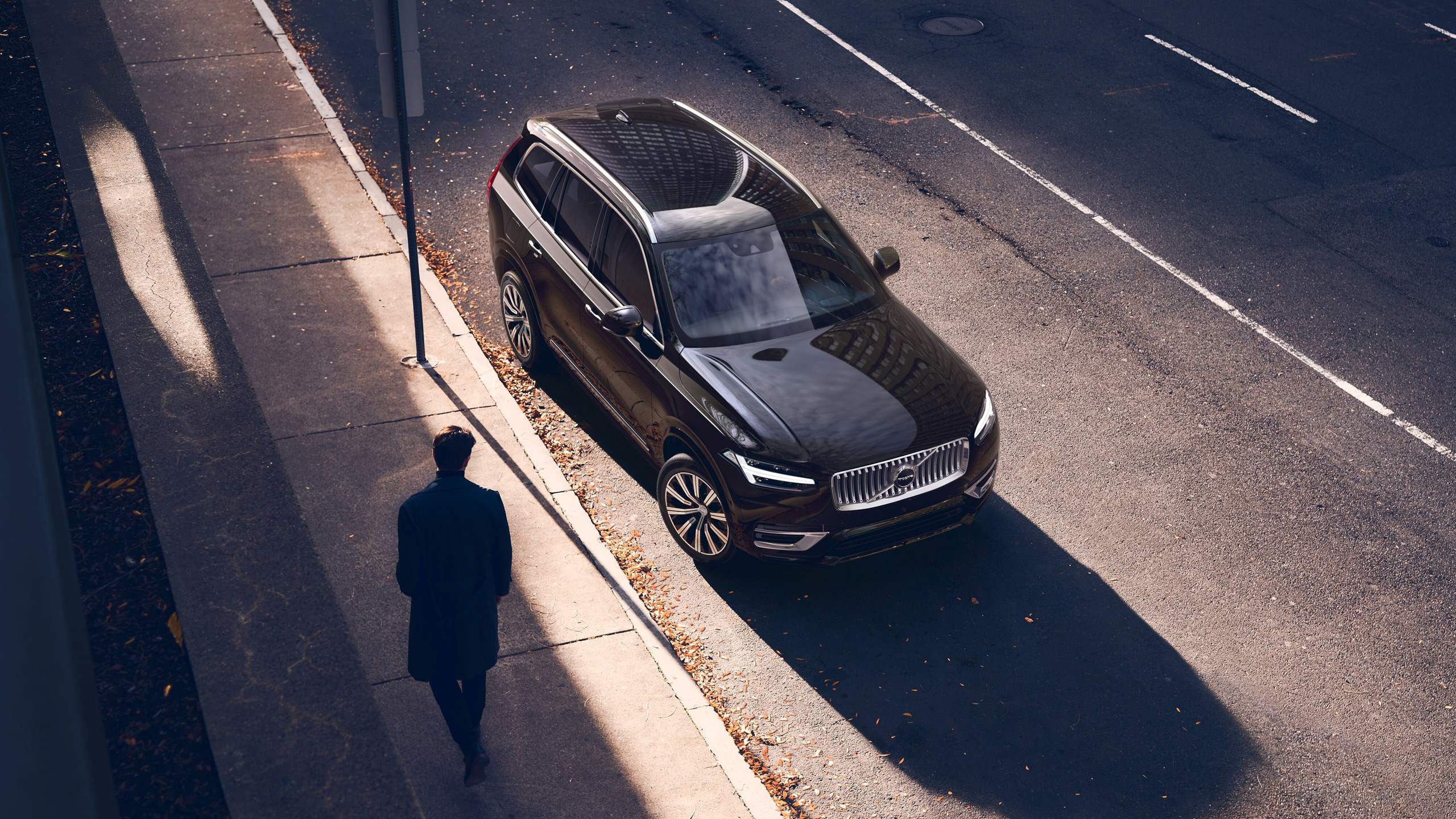 Vooraanzicht van bovenaf van de Volvo XC90 in Onyx Black, geparkeerd in een straat terwijl een man komt aanlopen, de zon staat laag en werpt lange schaduwen.