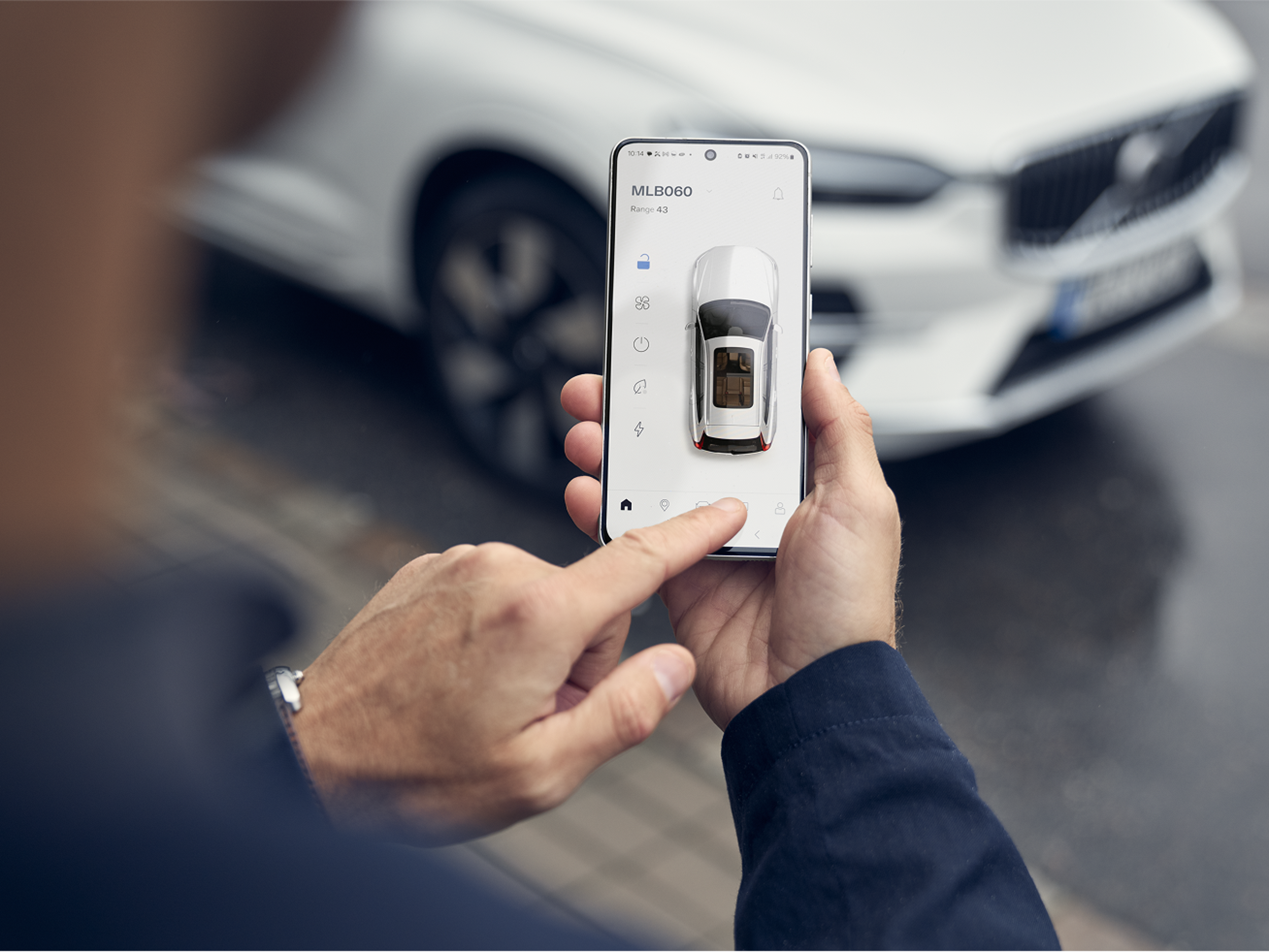 Persoon die de Volvo Cars app op een smartphone gebruikt om toegang te krijgen tot externe functies van het voertuig, met een witte Volvo zichtbaar op de achtergrond.