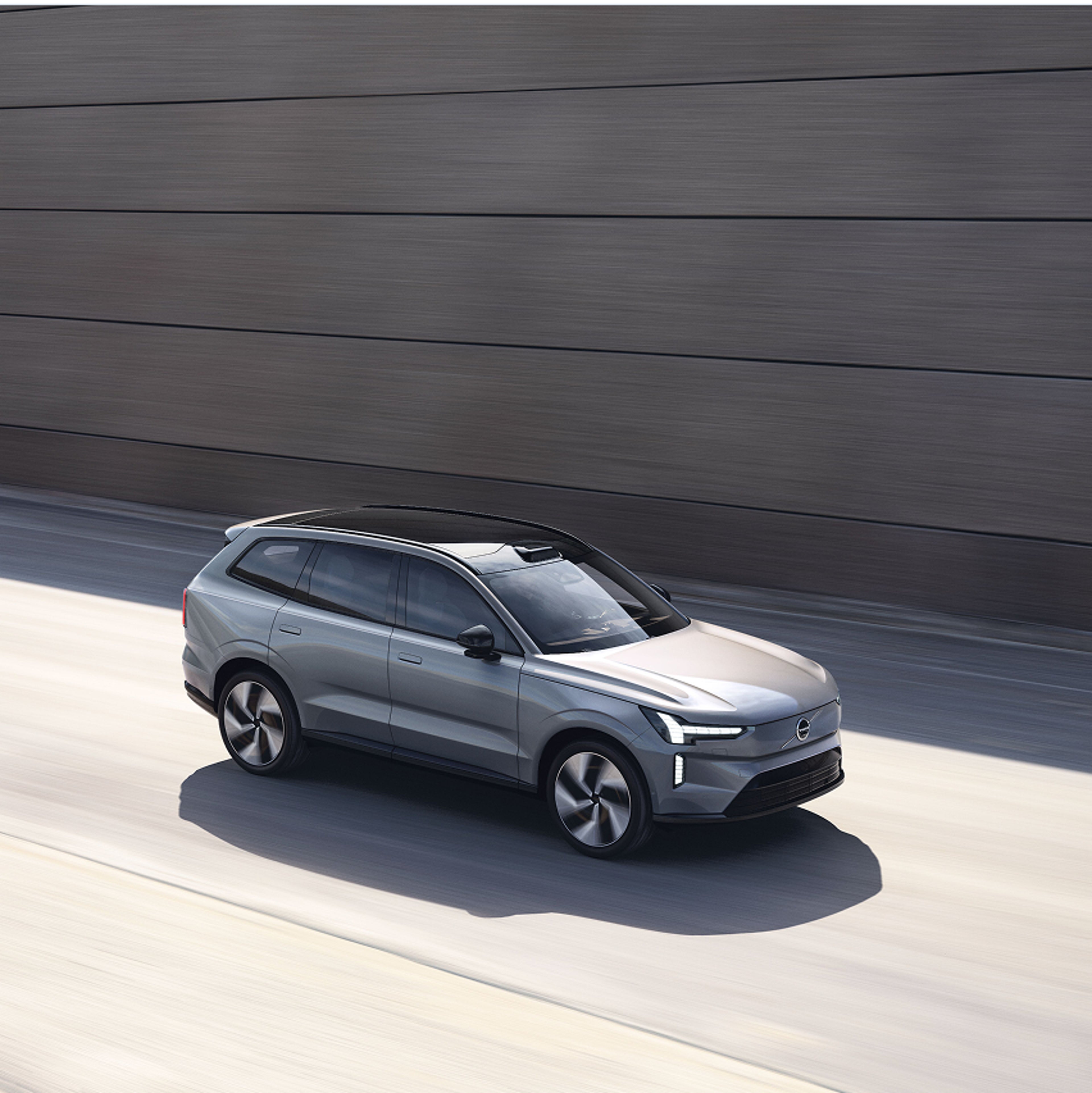 Šedé Volvo EX90 jedoucí po sluncem zalité silnici vedle moderní betonové zdi.