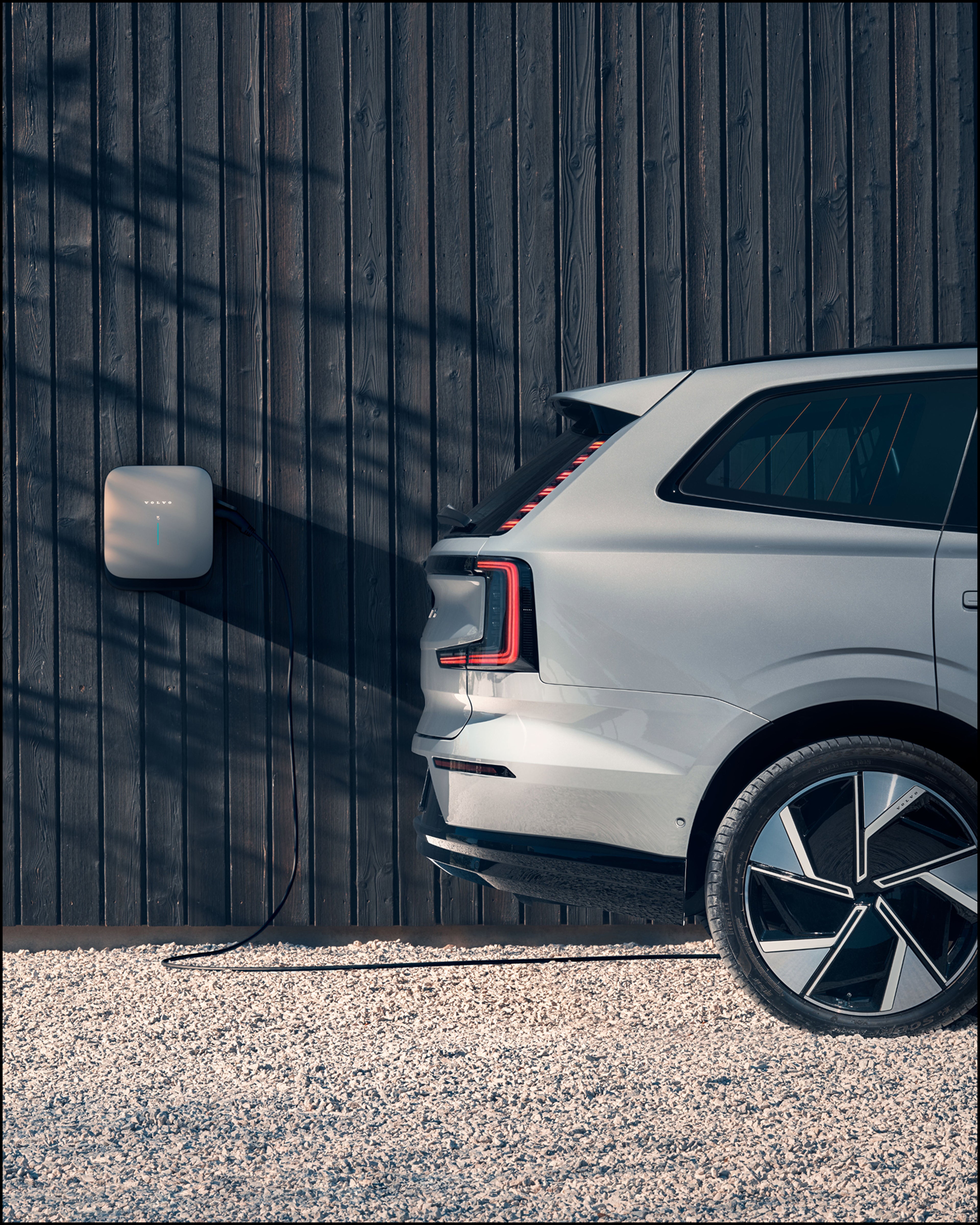 Side view of a silver Volvo electric SUV charging via a  wall-mounted Volvo wallbox on a dark wooden facade, in a sunlit outdoor setting.