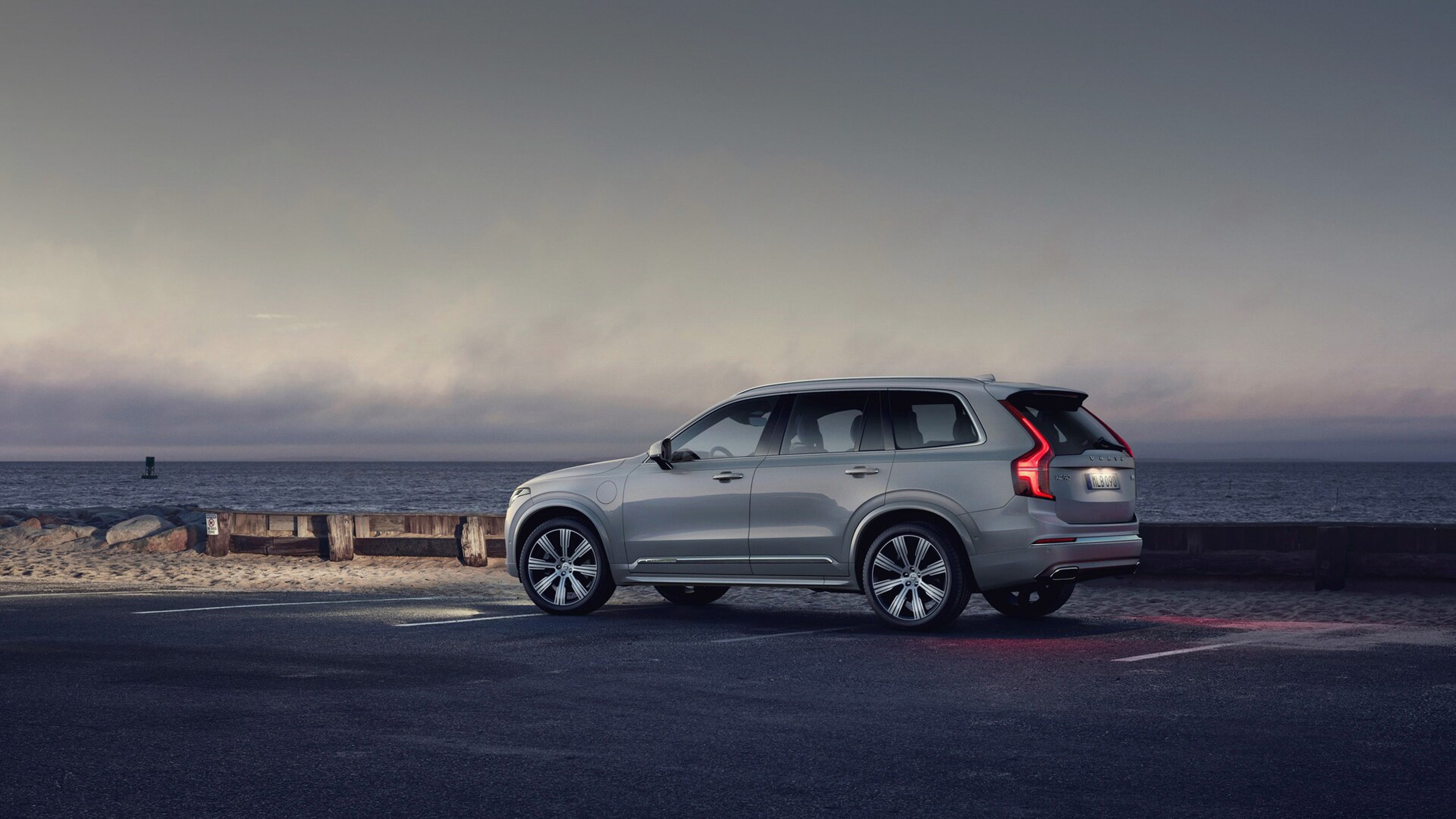 A side profile view of the Volvo XC90 in silver dawn.  Parked on the side of the road facing the sea with the car lights on.