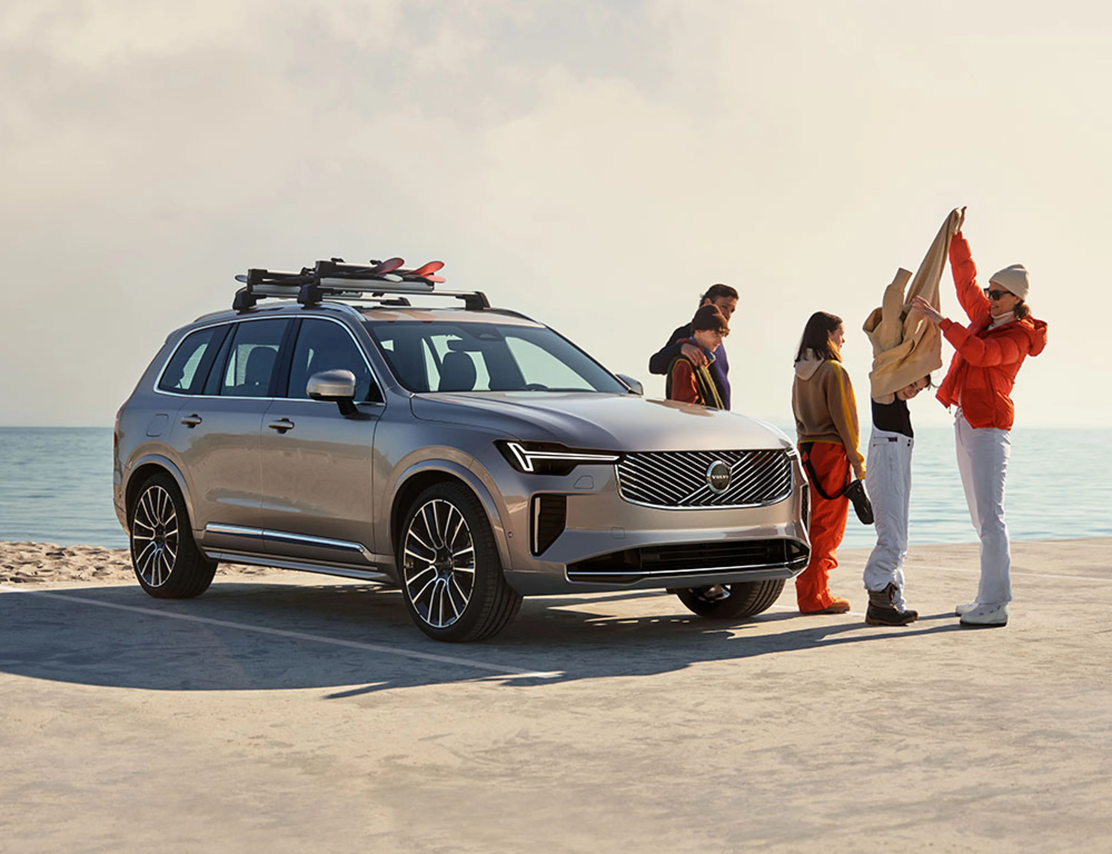 XC90 Argent Crépuscule vu de l’avant garé sur un parking près d’une plage avec une famille de 4 personnes