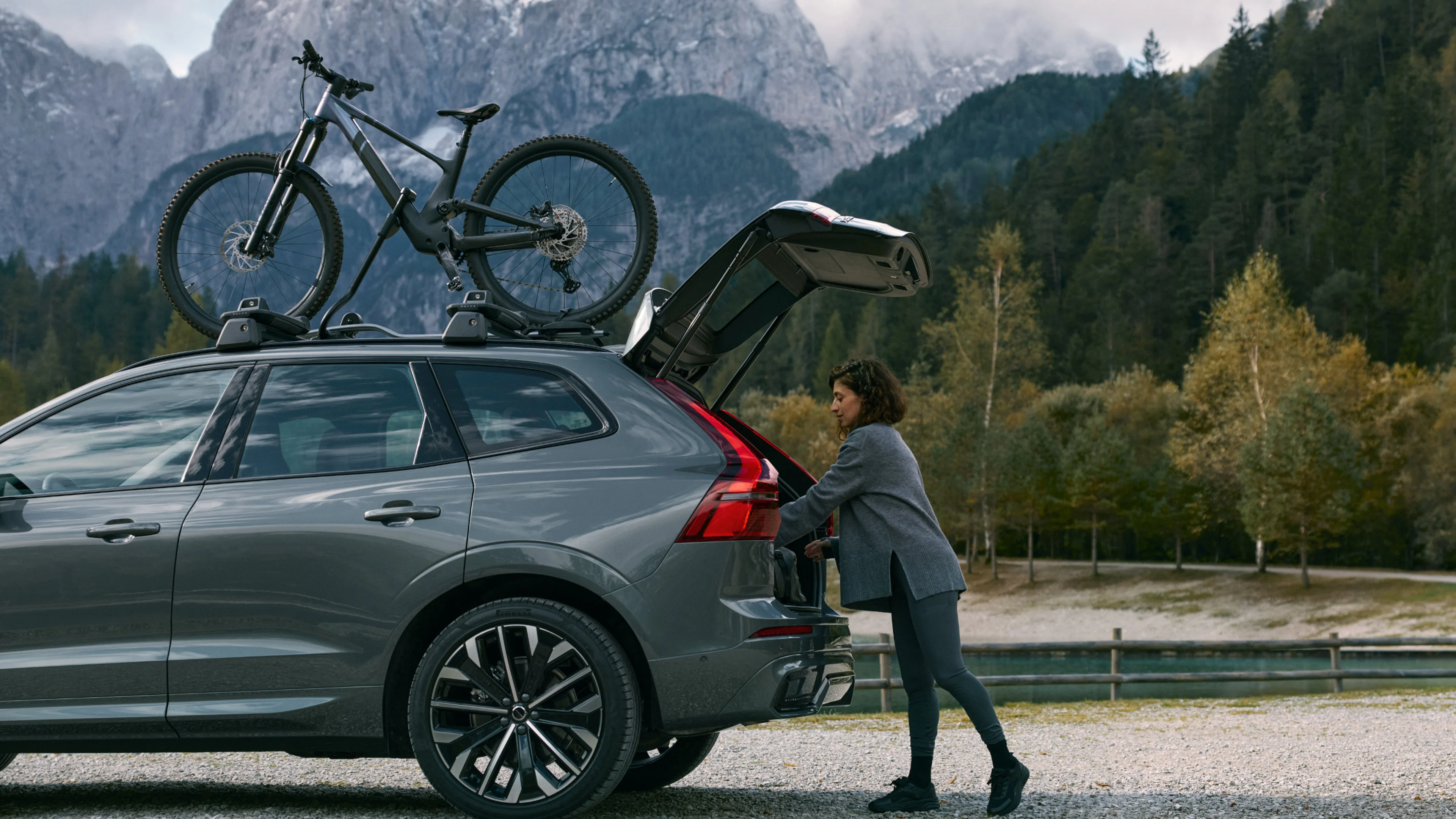 Ein Volvo XC60 mit Dachgepäckträger & montiertem Mountainbike steht vor einer Bergkulisse. An der geöffneten Heckklappe belädt eine Frau das Fahrzeuginnere.