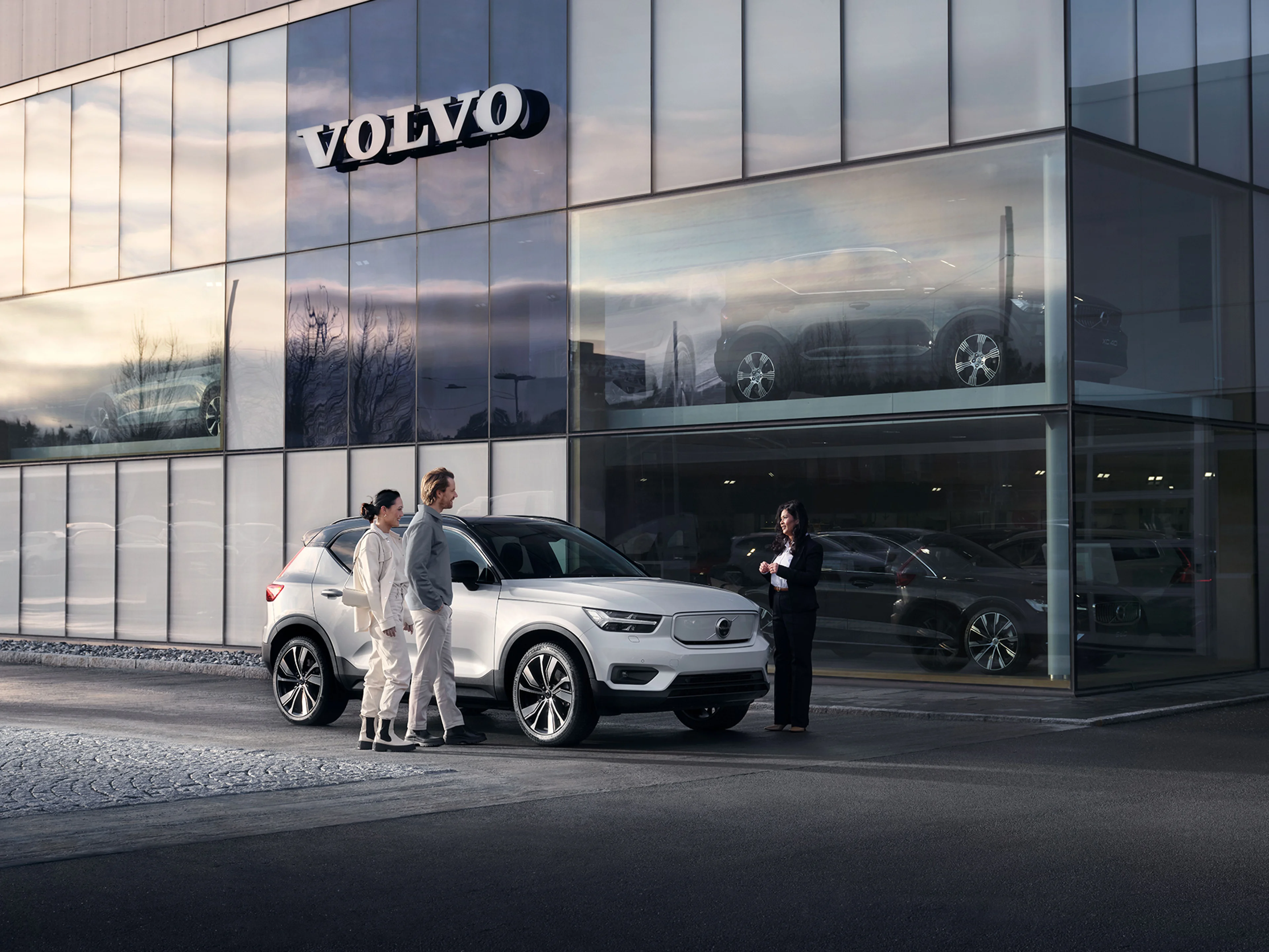 Ein Paar steht vor einem perlmuttfarbenen Volvo XC40, der widerrum vor einem Volvo Autohaus steht. Eine weibliche Verkaufsberaterin berät die Frau und den Mann.